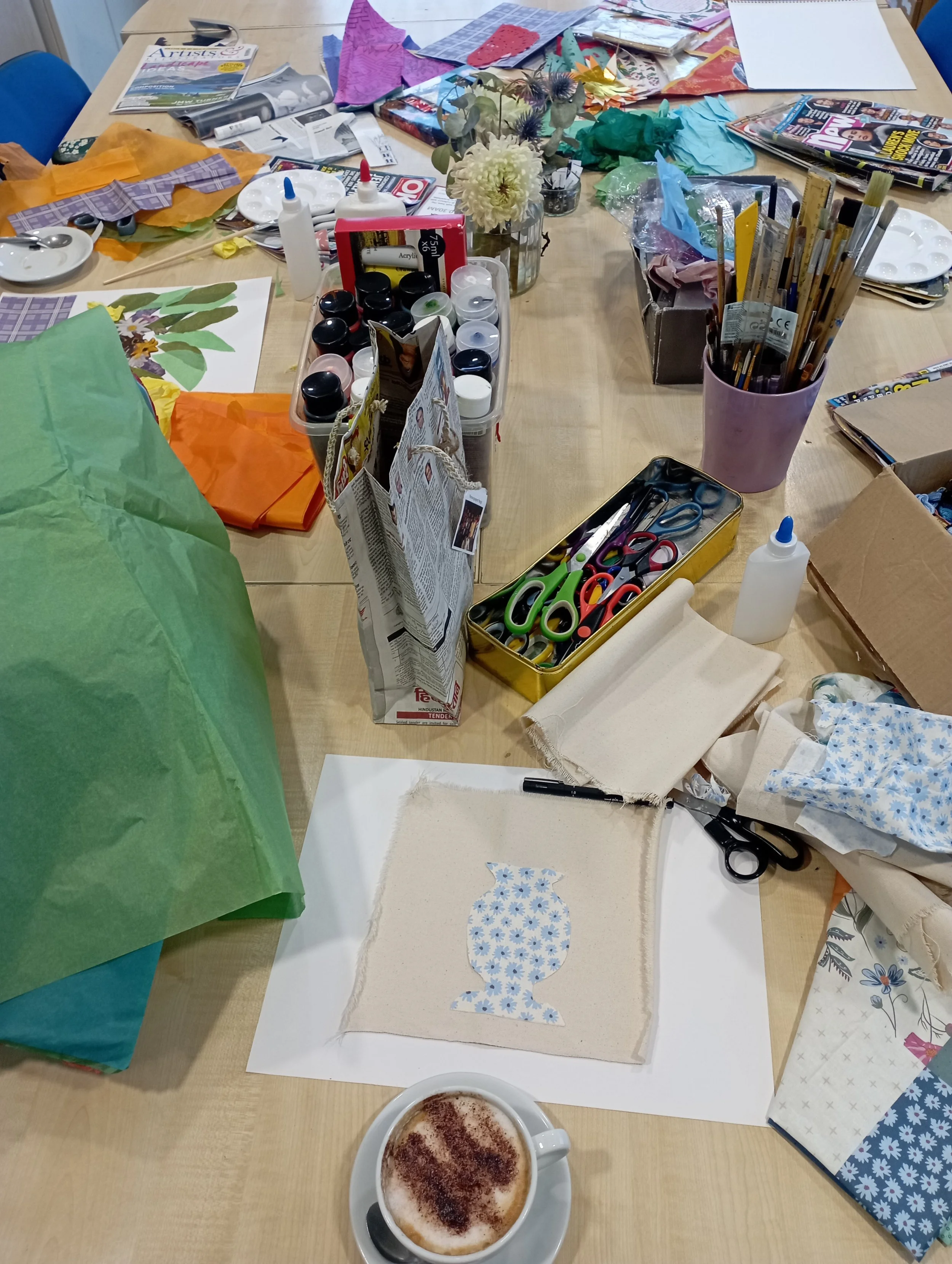 Arts and craft workspace with various materials, scissors, glue, fabric, paper, and a coffee cup on a table.