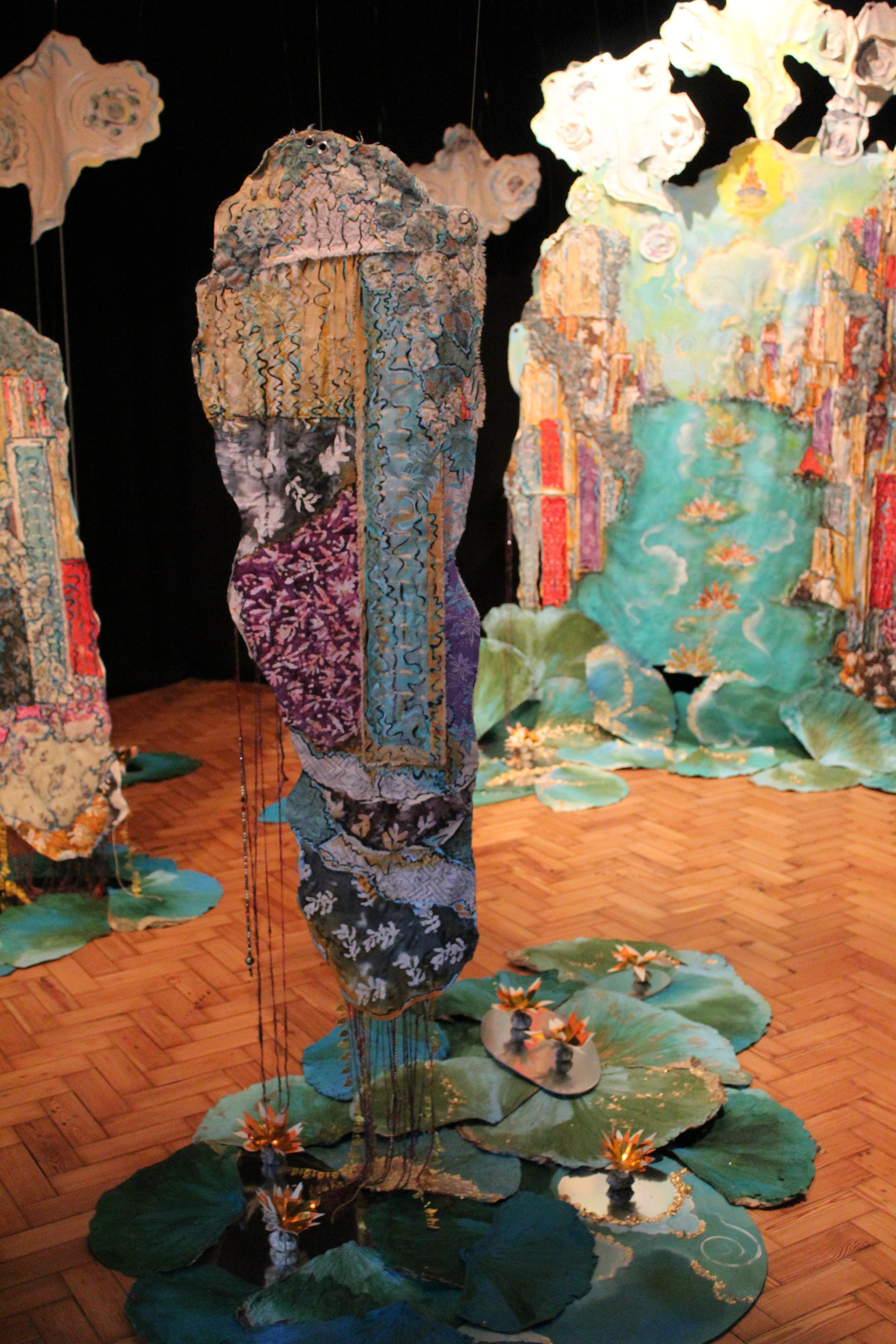 Mixed-media art installation featuring fabric sculptures resembling a moon face, surrounded by painted fabric panels and water lily decorations on a wooden floor.
