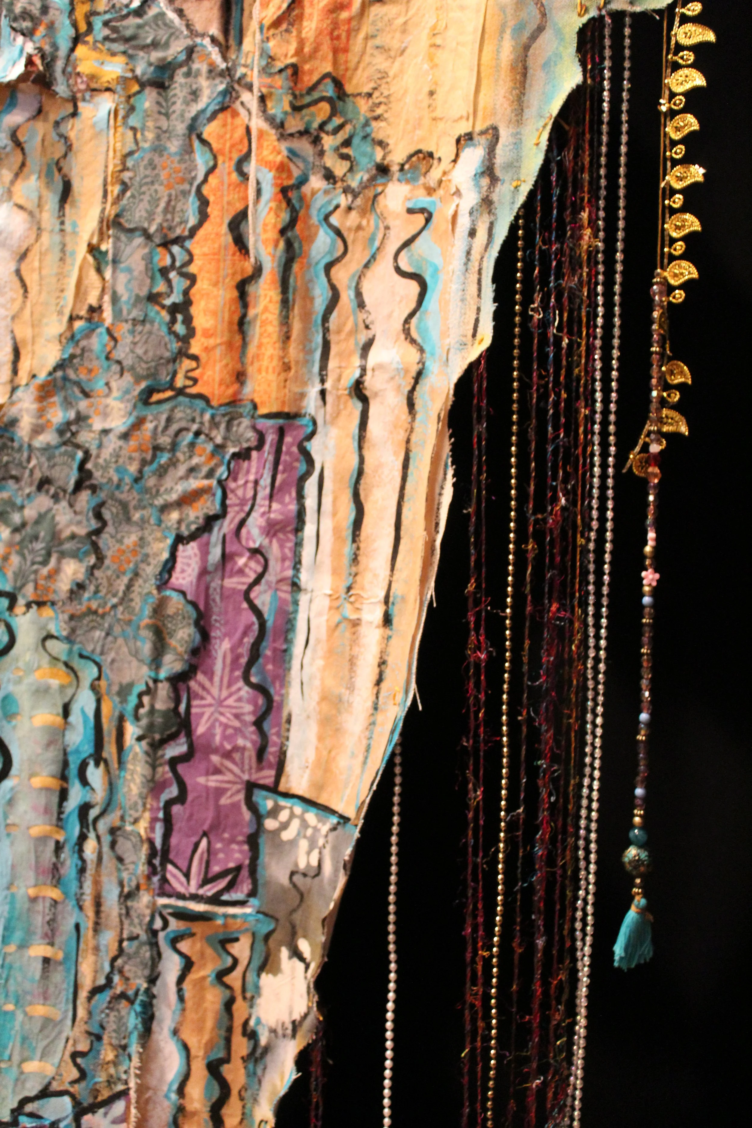 Close-up of a textured textile artwork with painted and stitched details on the left, and hanging layered beaded necklaces with tassels and metal embellishments on the right, against a black background.