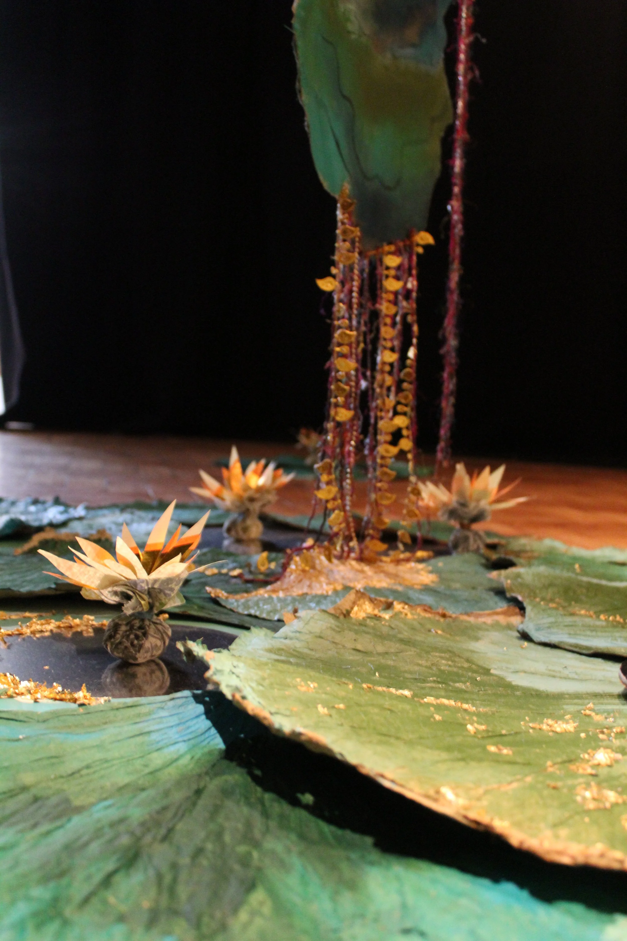 A close-up of an artistic arrangement featuring lotus-like paper flowers, large green leaves, and hanging strings of yellow and red beads, set against a dark background.