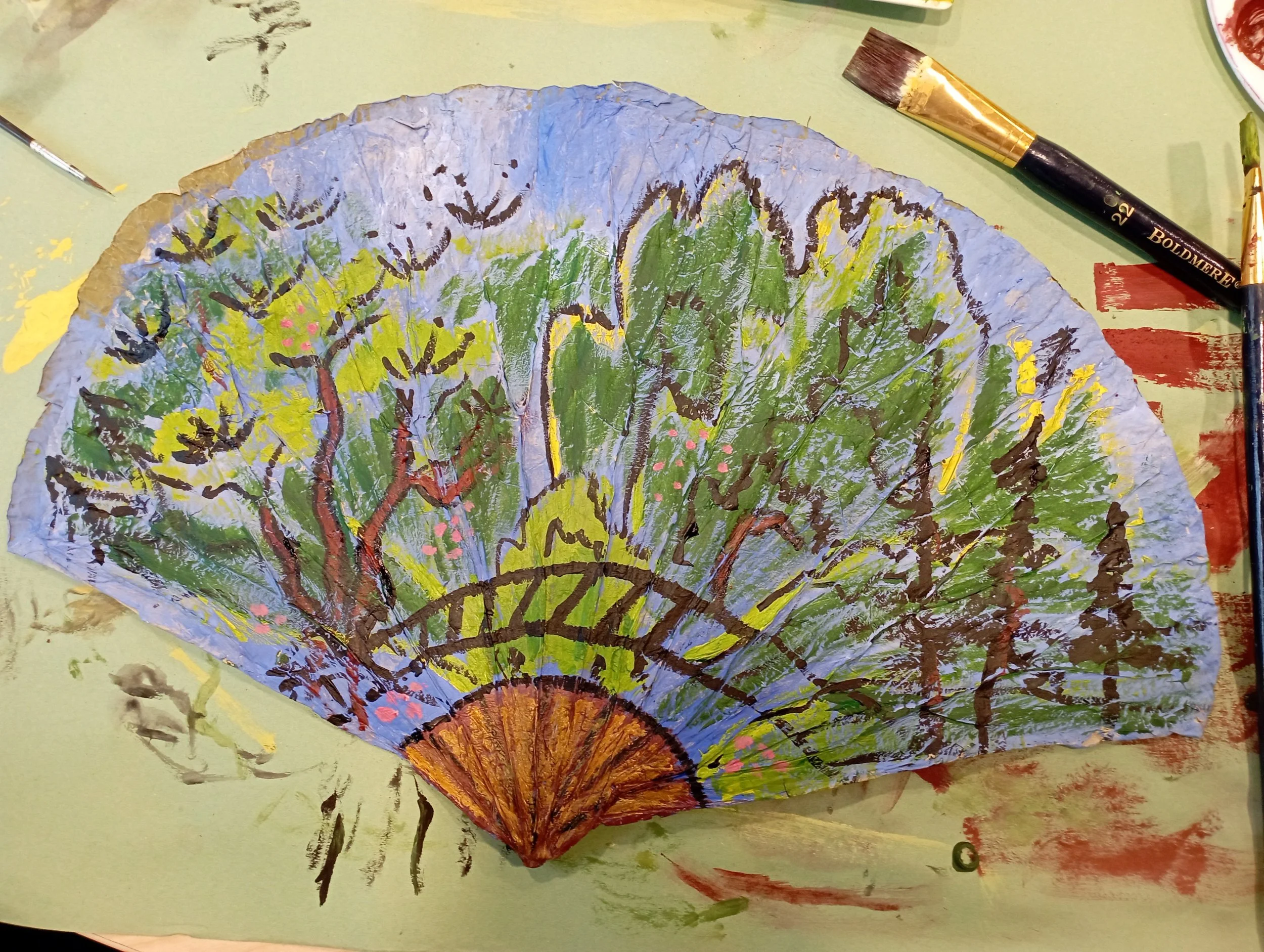 A painted paper fan showing a landscape with trees and a bridge, painted with green, blue, brown, pink, and yellow colors, on a table with paint brushes and other art supplies around it.