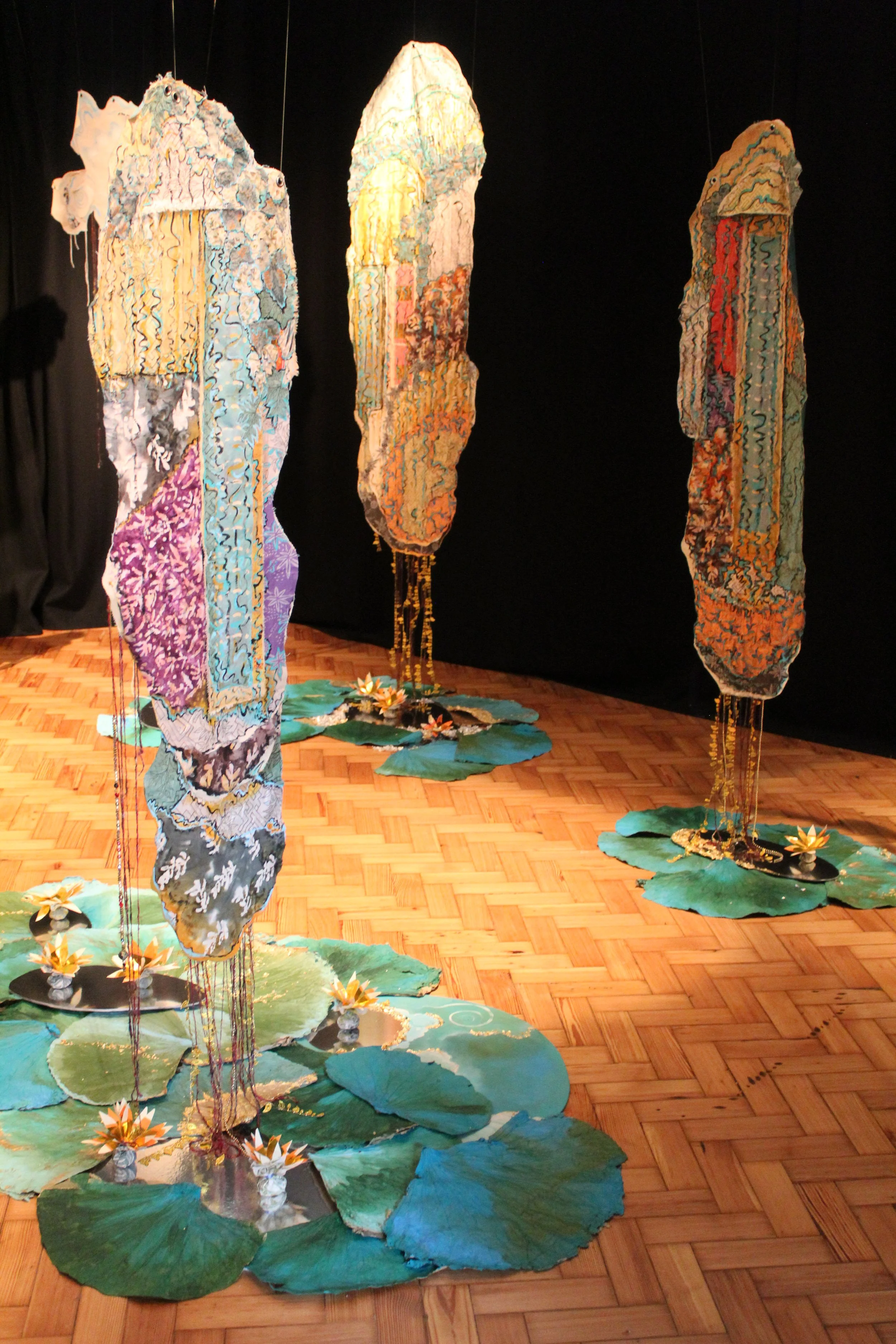 Three fabric art installations resembling abstract trees with lily pad bases, displayed in a gallery with a wooden floor.