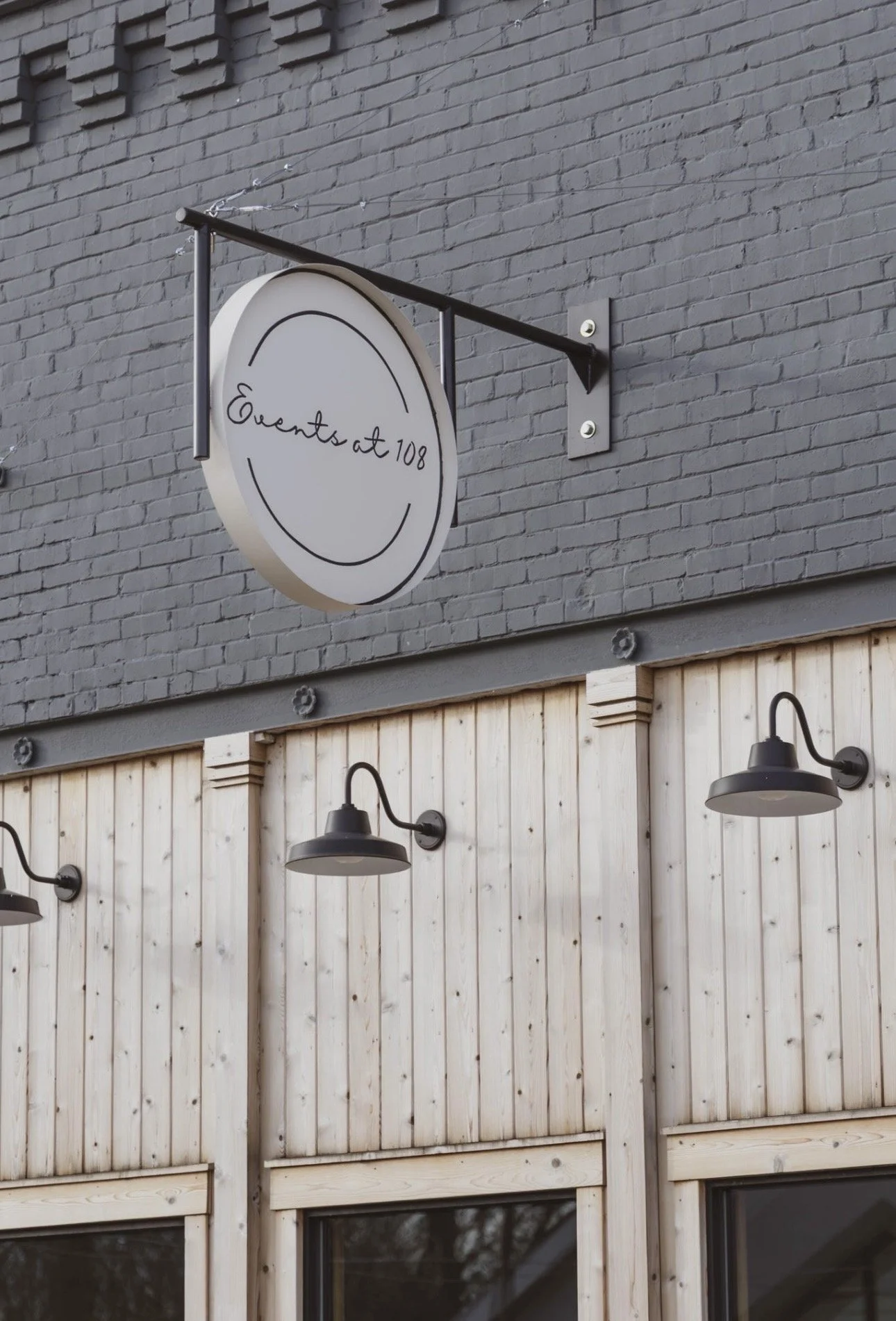 Sign for Events at 108 restaurant or event space mounted on a gray brick wall above a wooden fence with outdoor wall lamps.