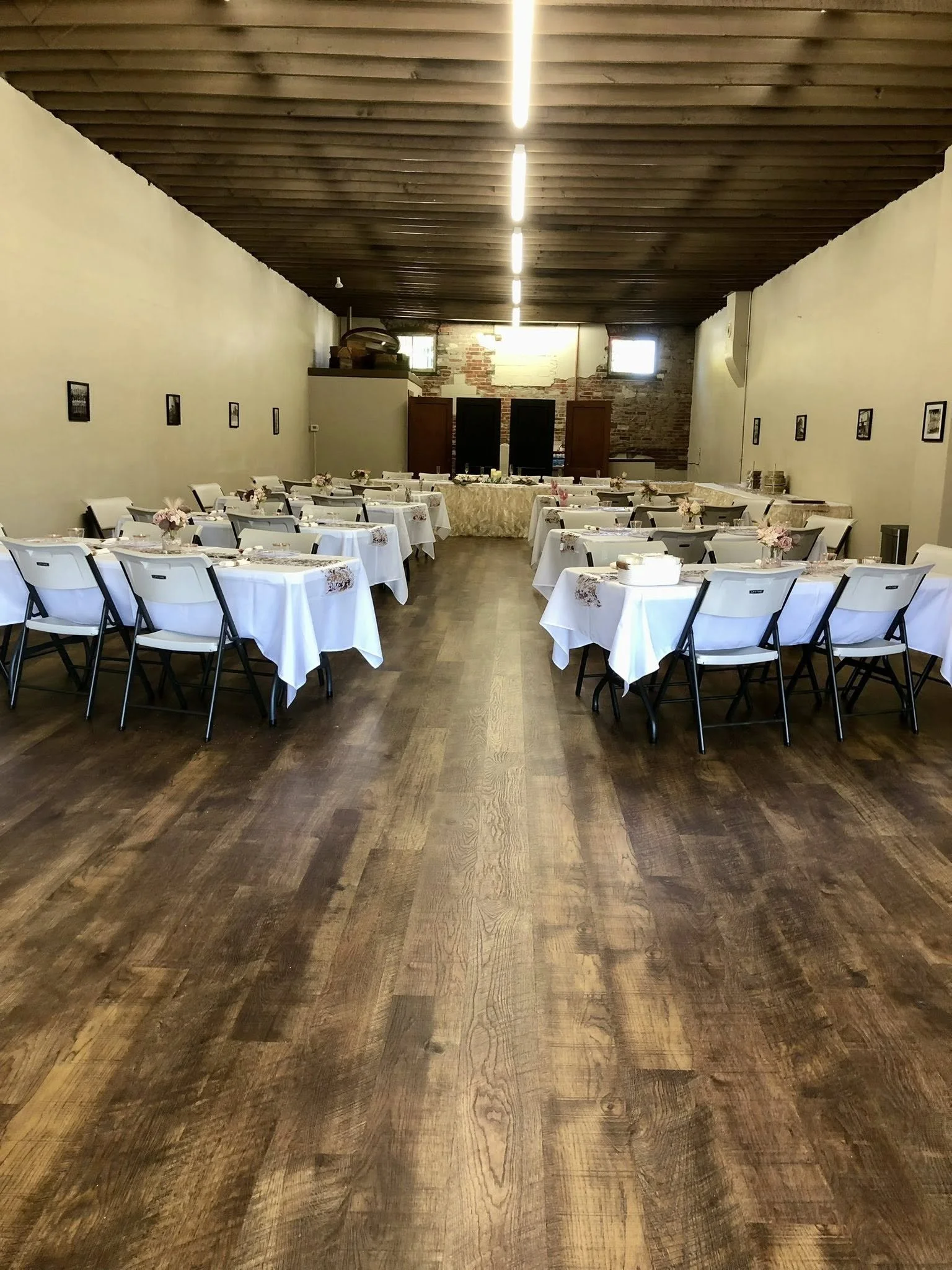 Empty banquet hall with white tablecloths, floral centerpieces, and chairs arranged around the tables, prepared for an event, with wooden floors and a wooden ceiling.