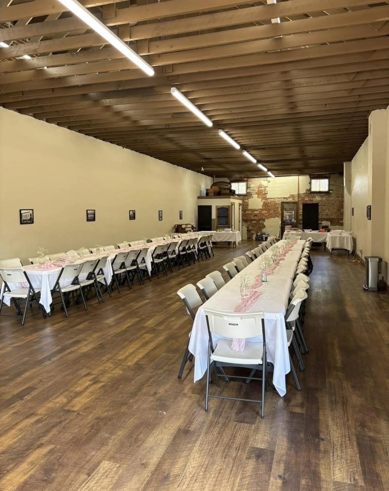 Long dining table set for a party or wedding or baby shower in a rustic hall with wood floors, white tablecloths, pink napkins, and small vases, with additional tables along the wall and a brick wall at the end.
