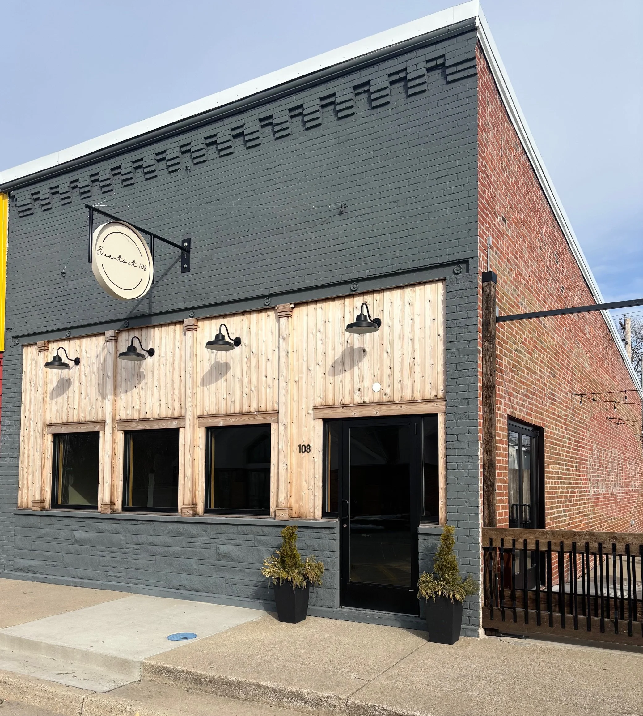 Exterior of a modern building with dark gray and brick walls, wooden paneling, and three window storefronts. Sign reads 'Event, at 108.' Potted plants are placed outside near the entrance, and black industrial-style lights are mounted above the windo
