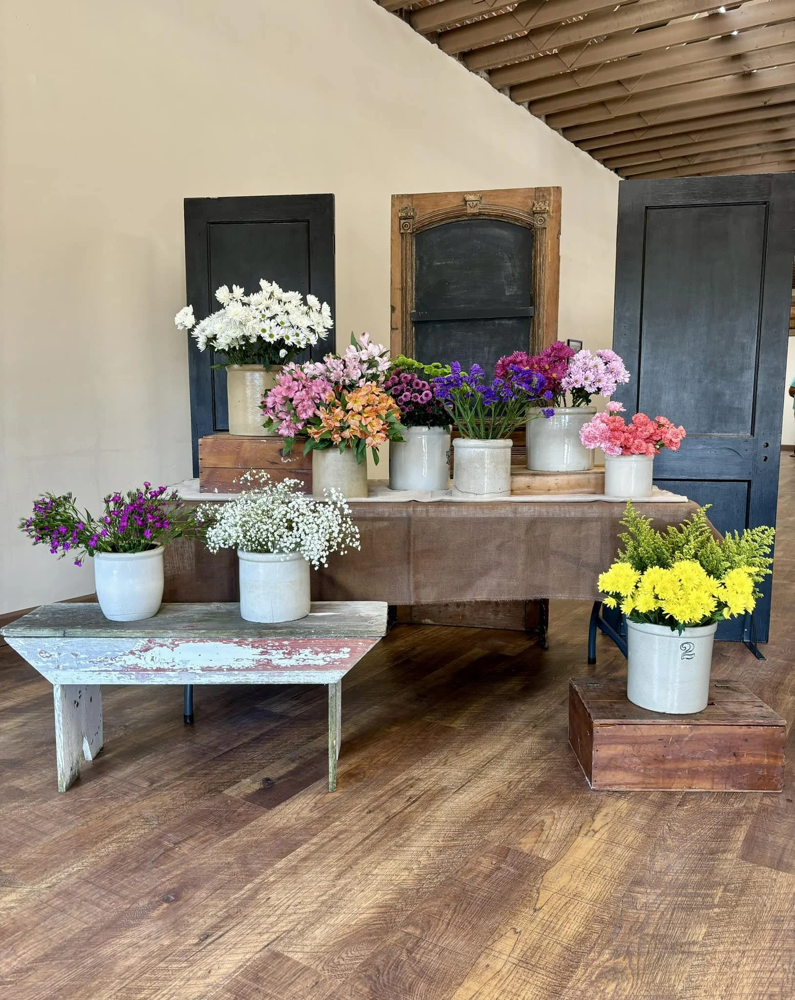 Various colorful flowers in white and beige pots arranged on a rustic table, with black chalkboards and window shutters as a backdrop.