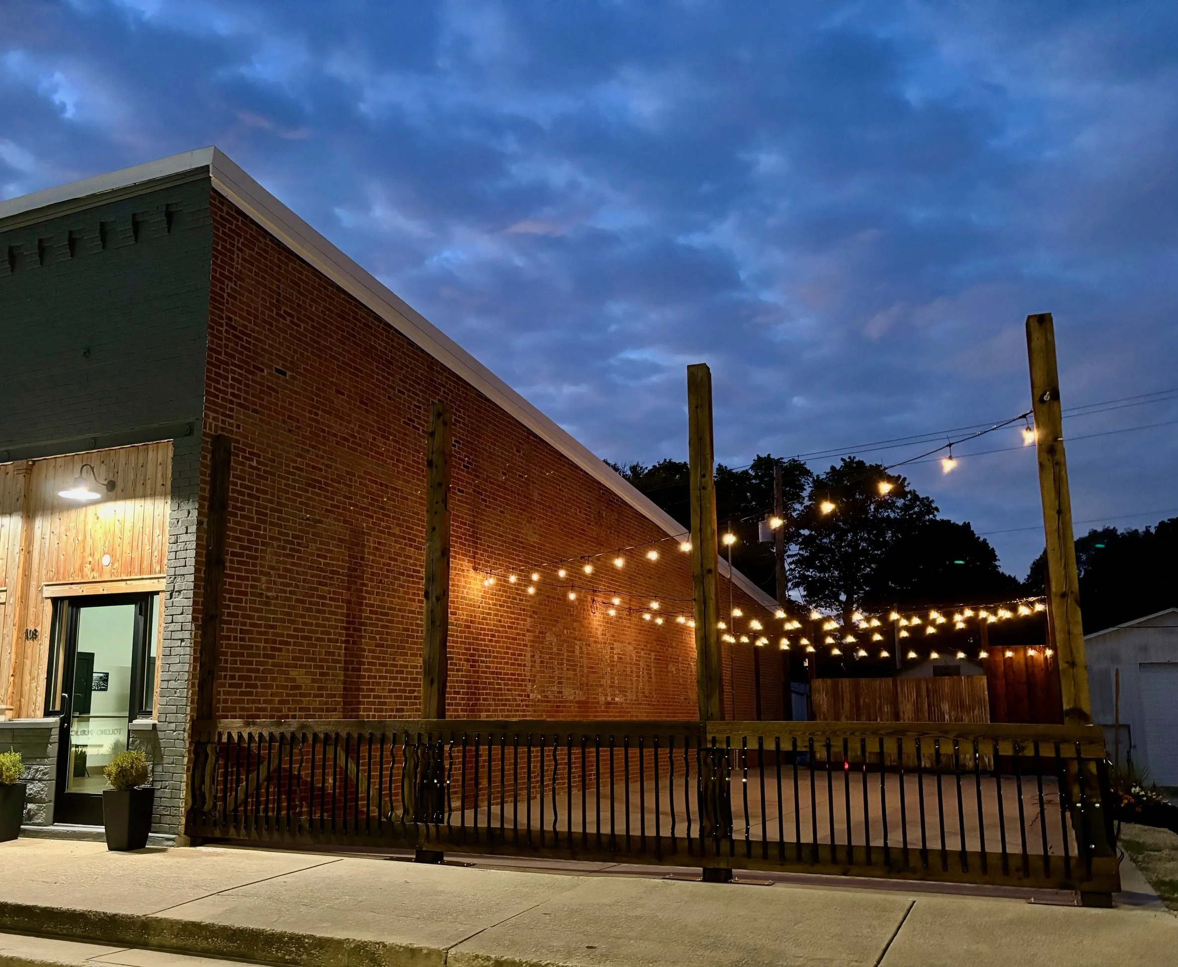 Nighttime view of Events at 108 with string lights hanging outside, a fenced patio area with wooden beams, and a cloudy sky in the background.