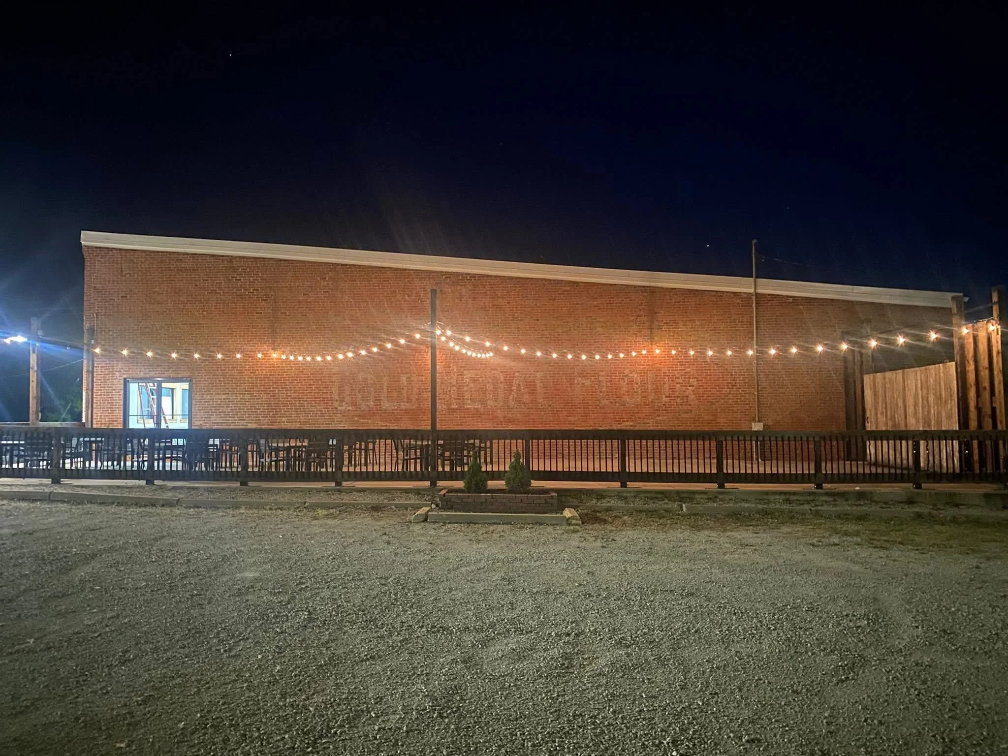 Night view of a brick building with a string of hanging lights and a small open window, surrounded by a wooden fence and a gravel parking lot.