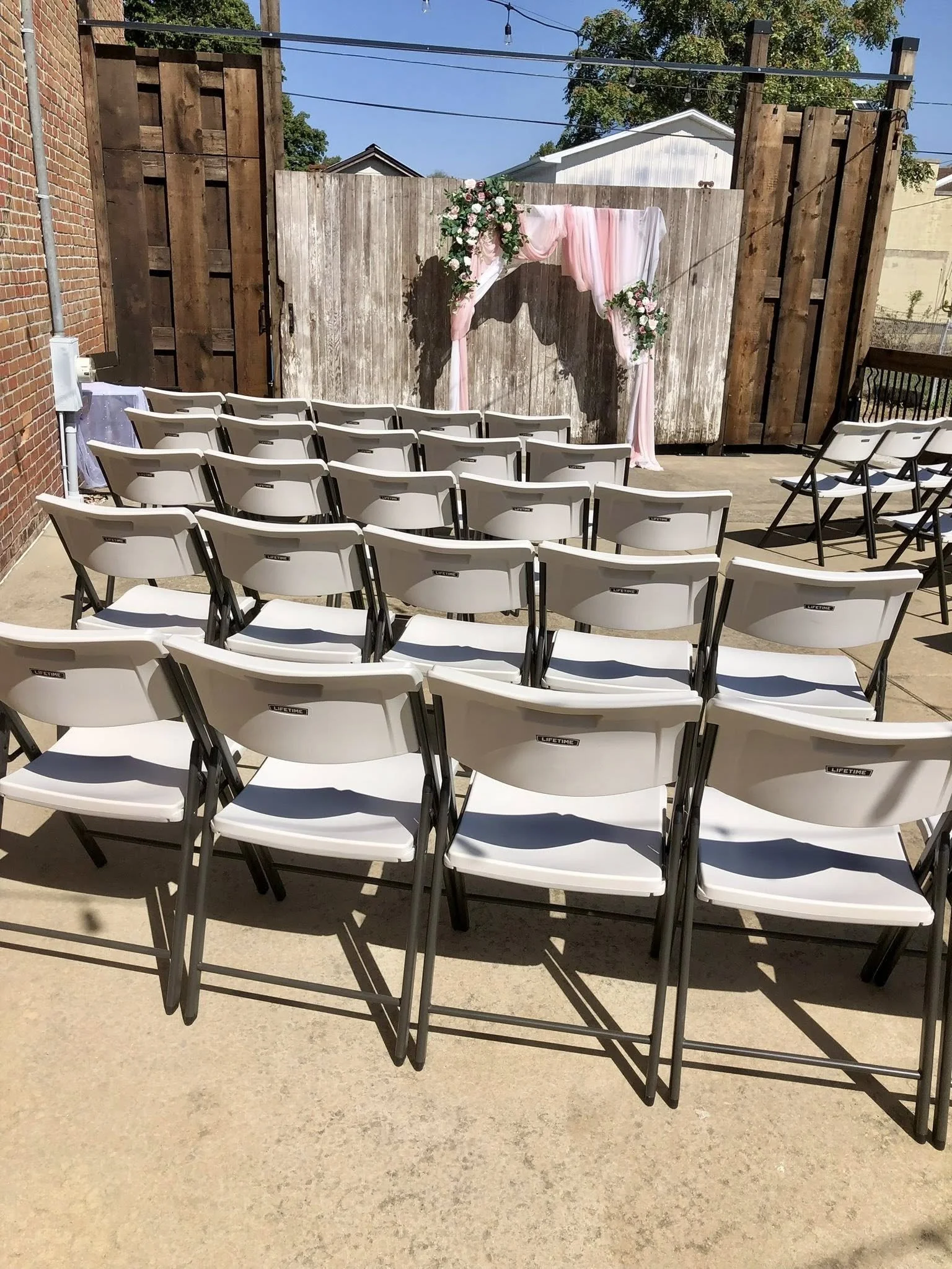 Outdoor wedding ceremony setup with white folding chairs arranged facing a decorated archway with pink and white fabric and floral arrangements against a wooden fence.