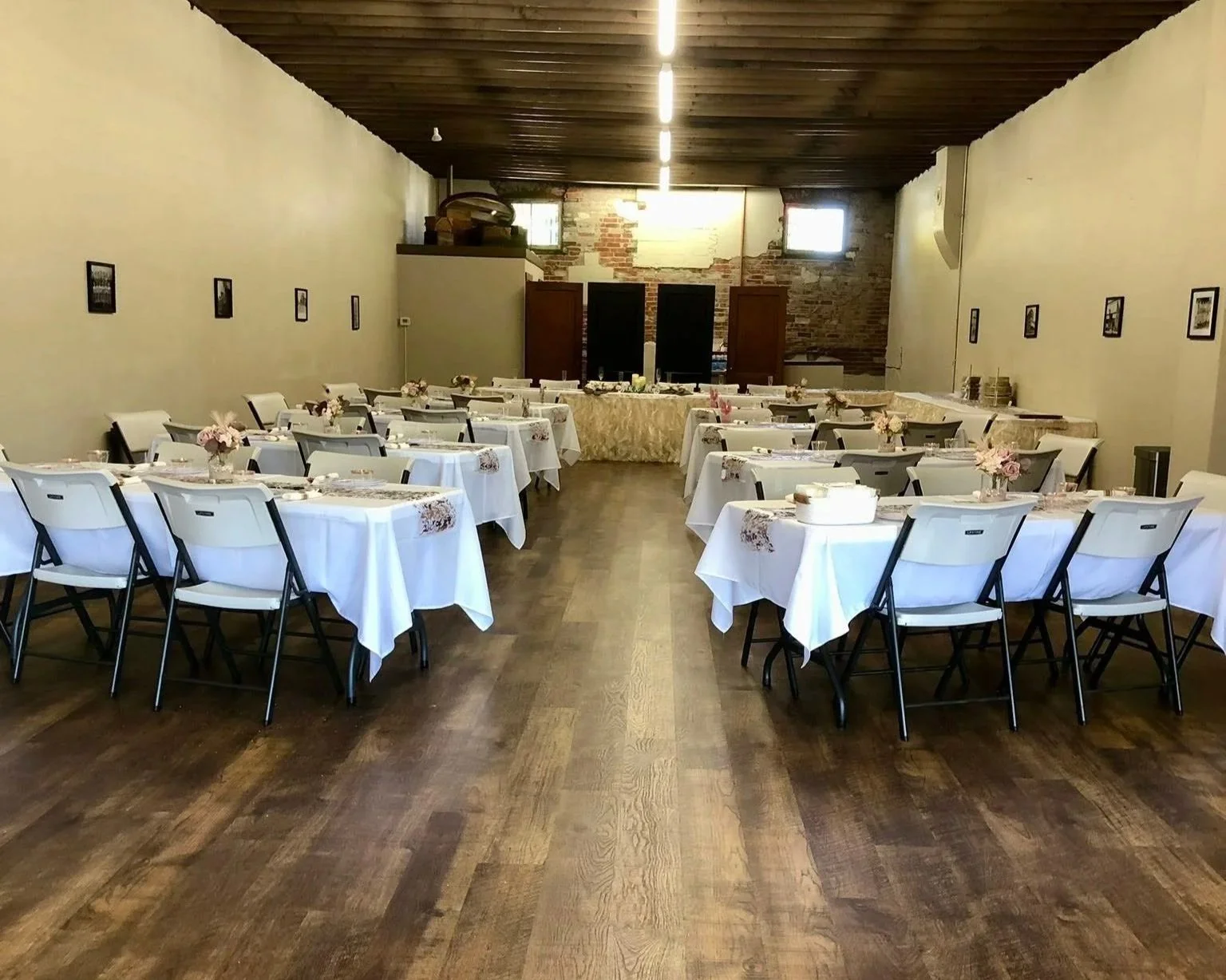 A banquet hall with round tables covered in white tablecloths, each decorated with small flower arrangements, set for a formal event. The hall has a wooden ceiling, brick walls at the back.