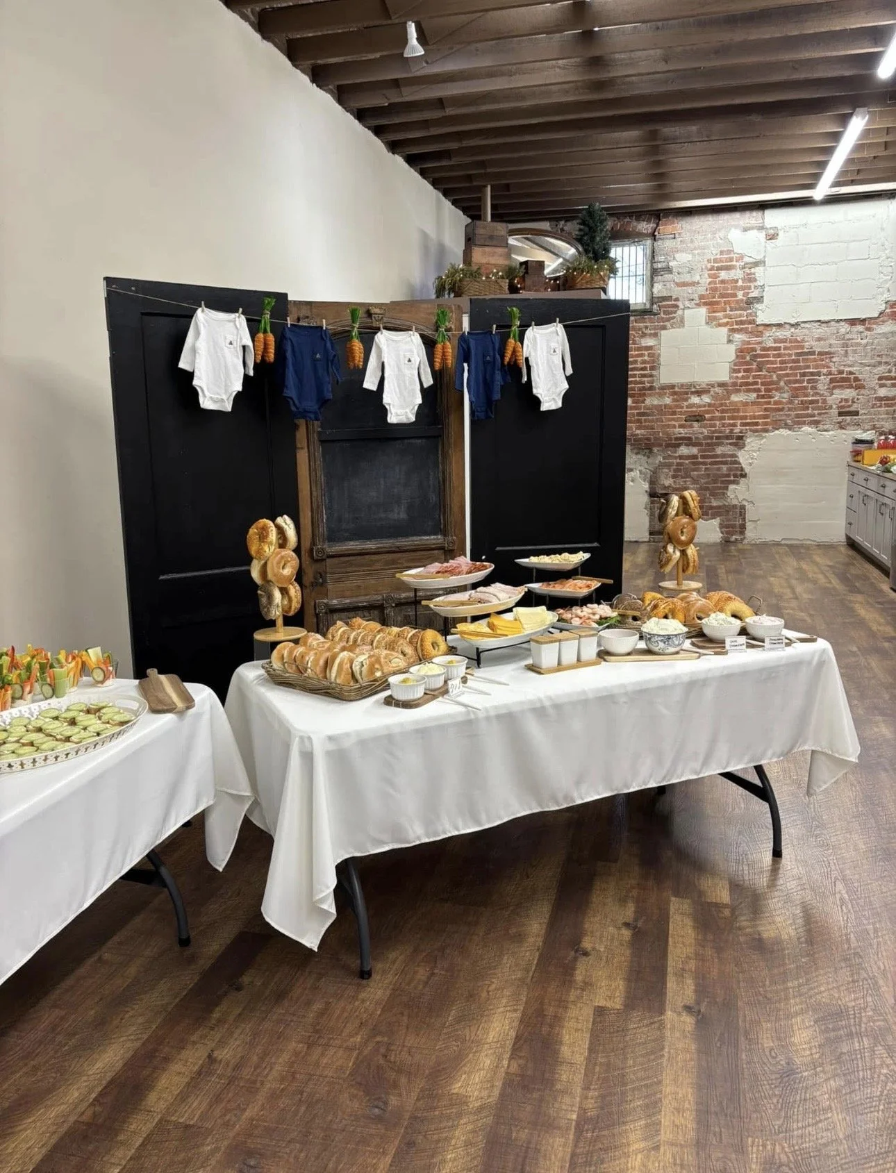 Breakfast buffet with bagels, sliced vegetables, and cold cuts, decorated for a holiday with baby clothes and hanging mini carrots on a table with a white tablecloth, in a rustic brick and wood decorated room.