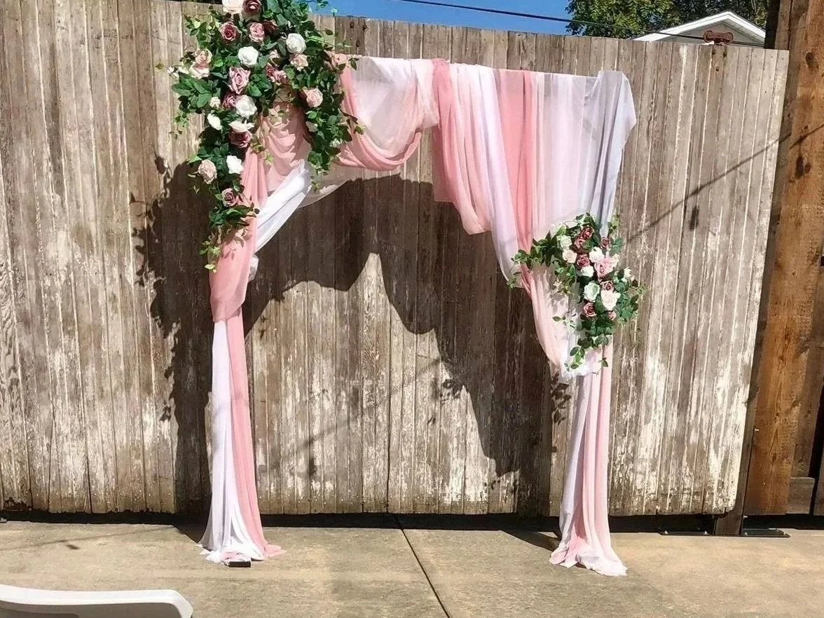 A decorative wedding arch with pink and white fabric and floral arrangements with pink and white roses against a wooden fence.
