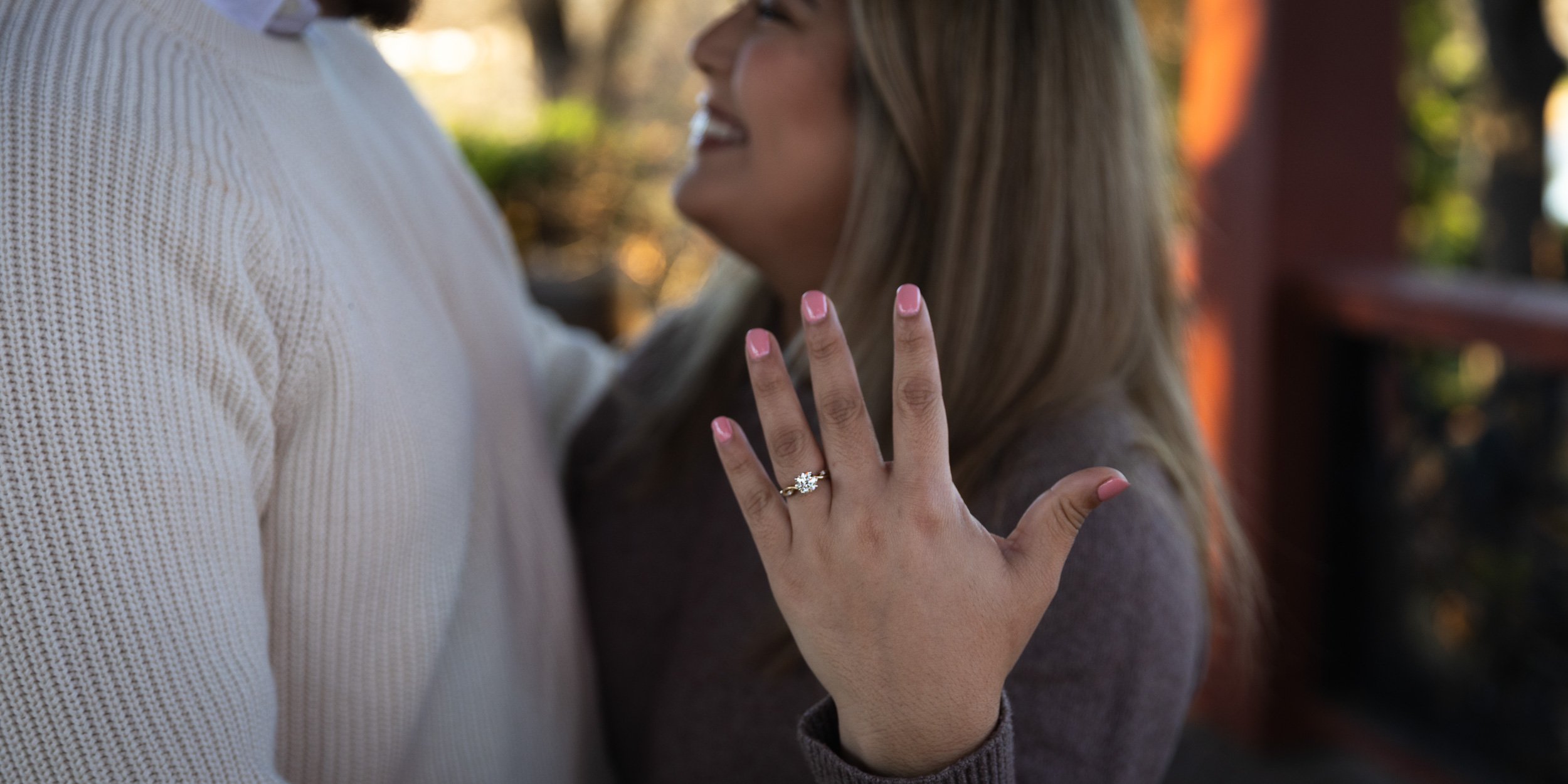 Close-up of a woman's hand showing an engagement ring with a large gemstone, as she touches a man's chest. The woman has long, blonde hair and is smiling. The man is wearing a white knitted sweater. The background is blurred with warm sunlight.