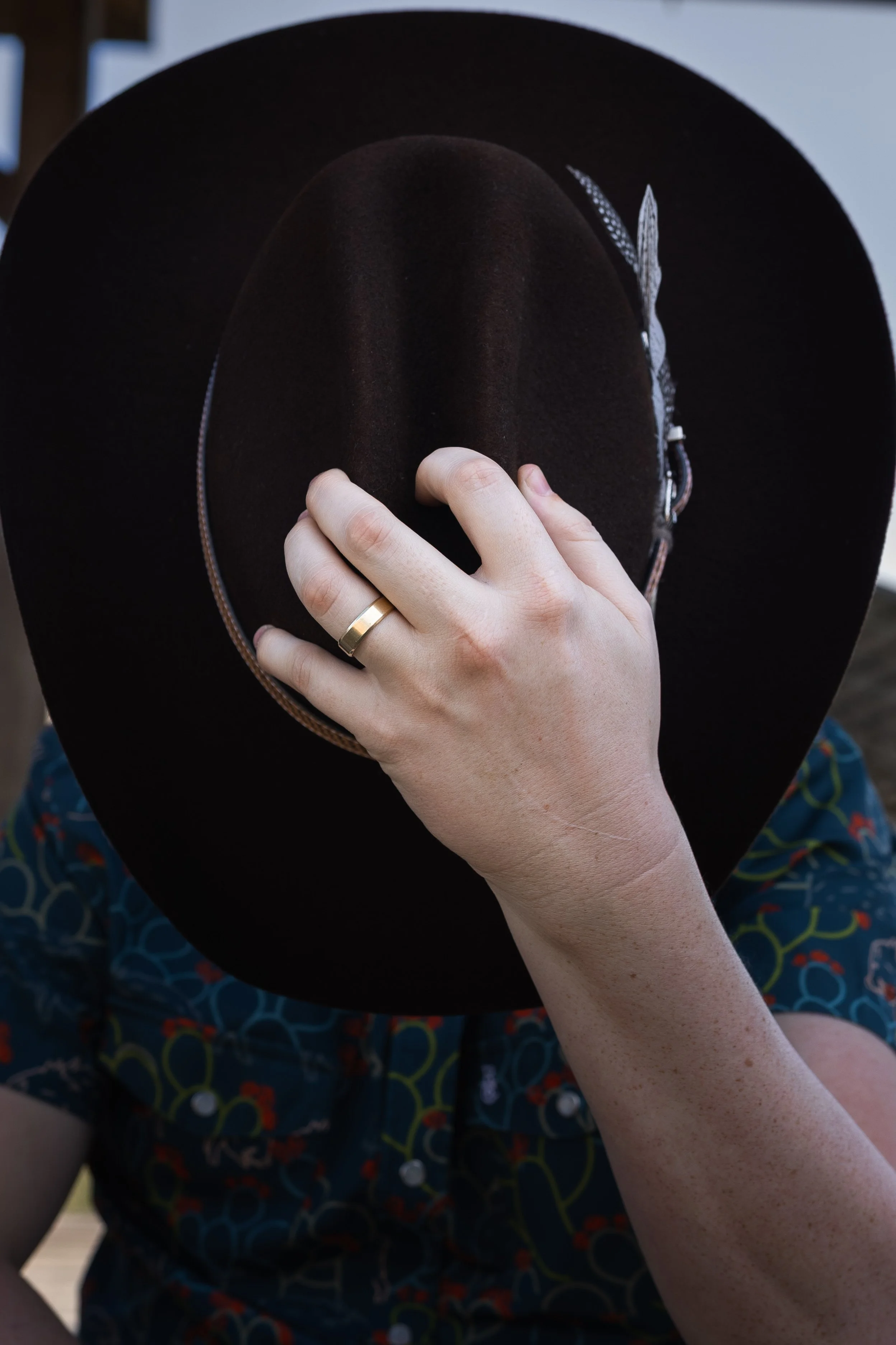 Person holding a brown felt hat with a decorative band, wearing a ring on their finger, and dressed in a colorful patterned shirt.