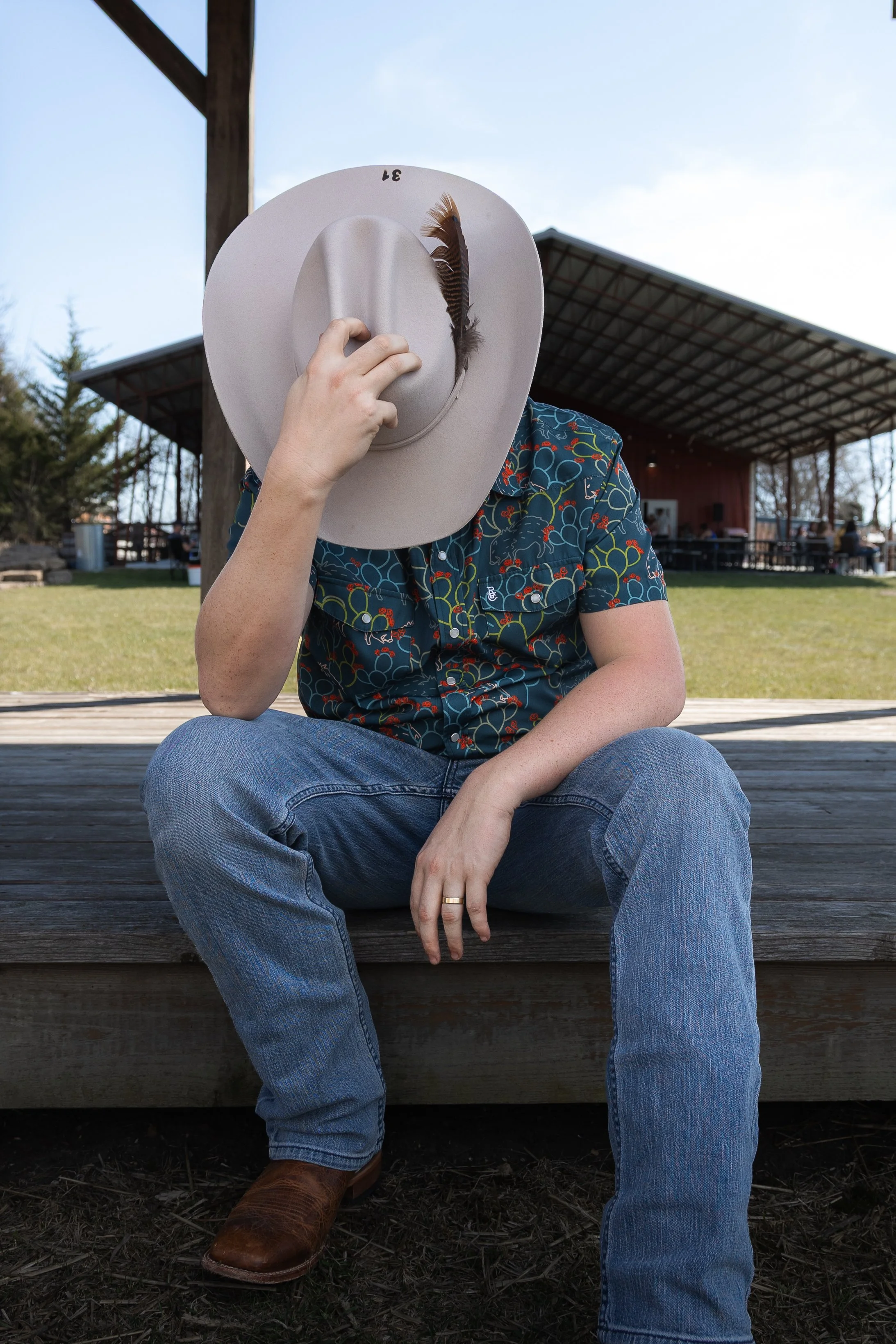 A person sitting outdoors on a wooden surface, wearing blue jeans, brown cowboy boots, a patterned shirt, and a wide-brimmed hat that covers their face, holding the hat's brim with their left hand, with a building and trees in the background.