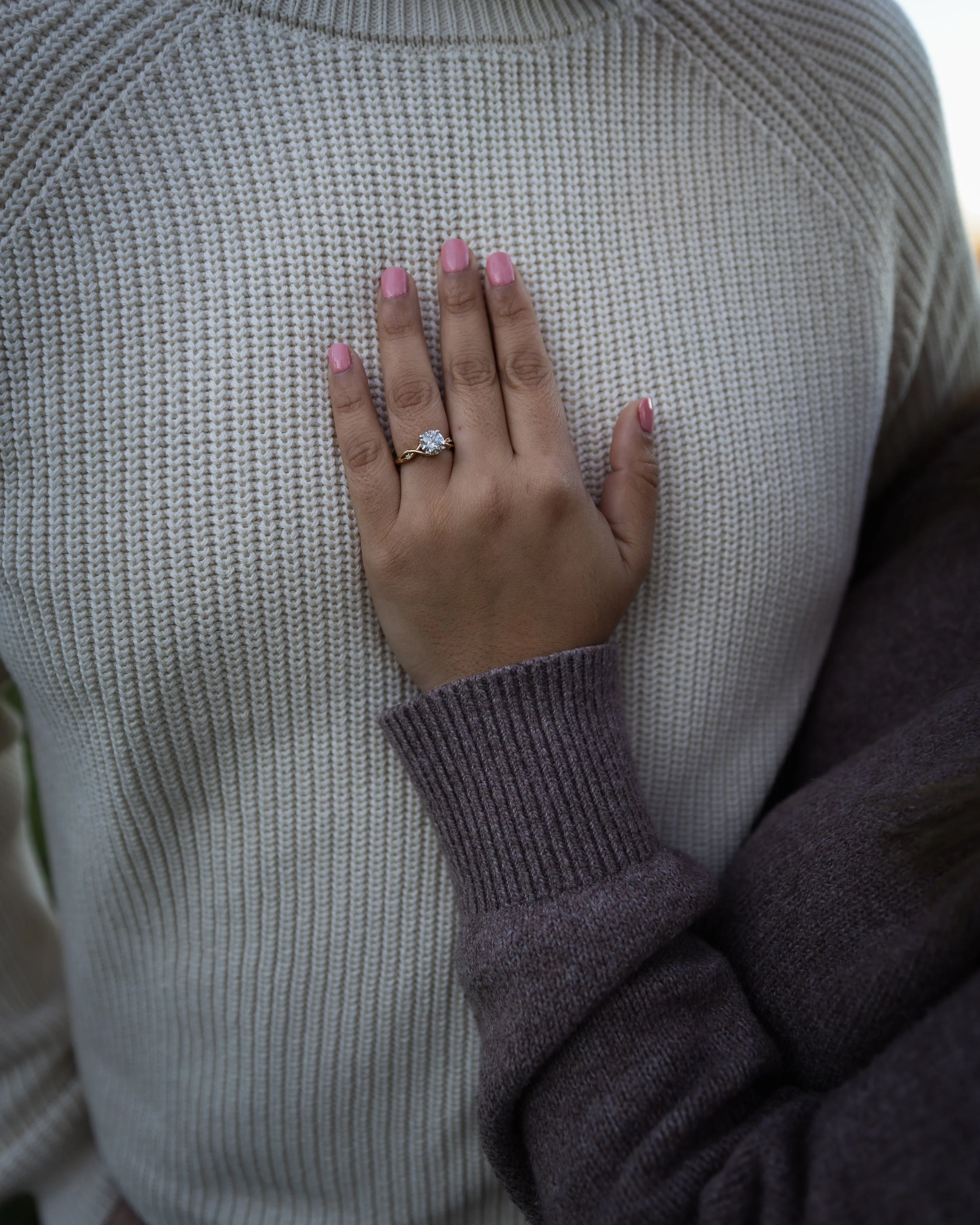 Close-up of a hand with pink painted nails wearing a diamond ring, resting on a person wearing a cream-colored knit sweater and a grey and purple sleeve.