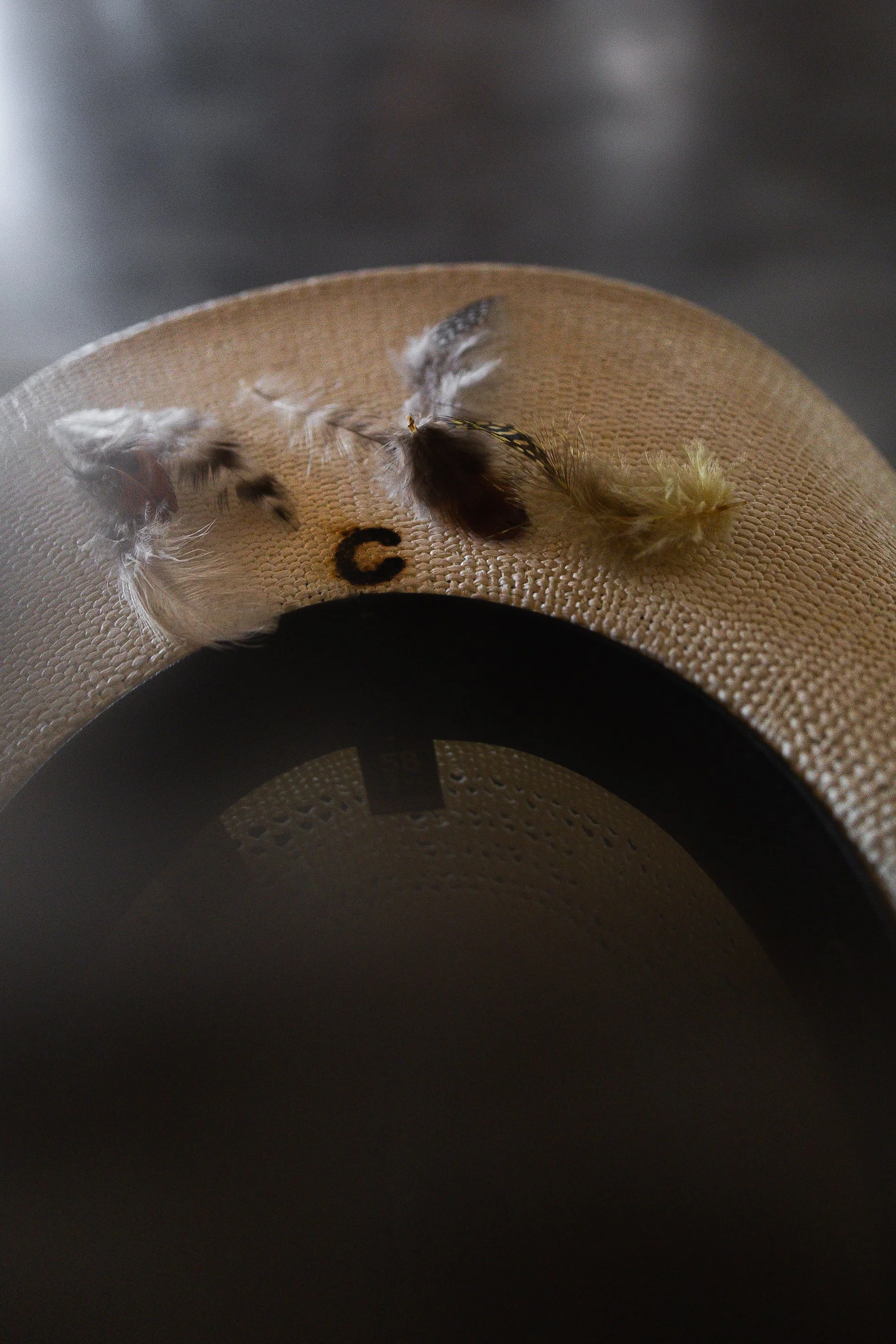Close-up of a beige fabric hat with small feathers attached, resting on a dark surface with a blurred background.