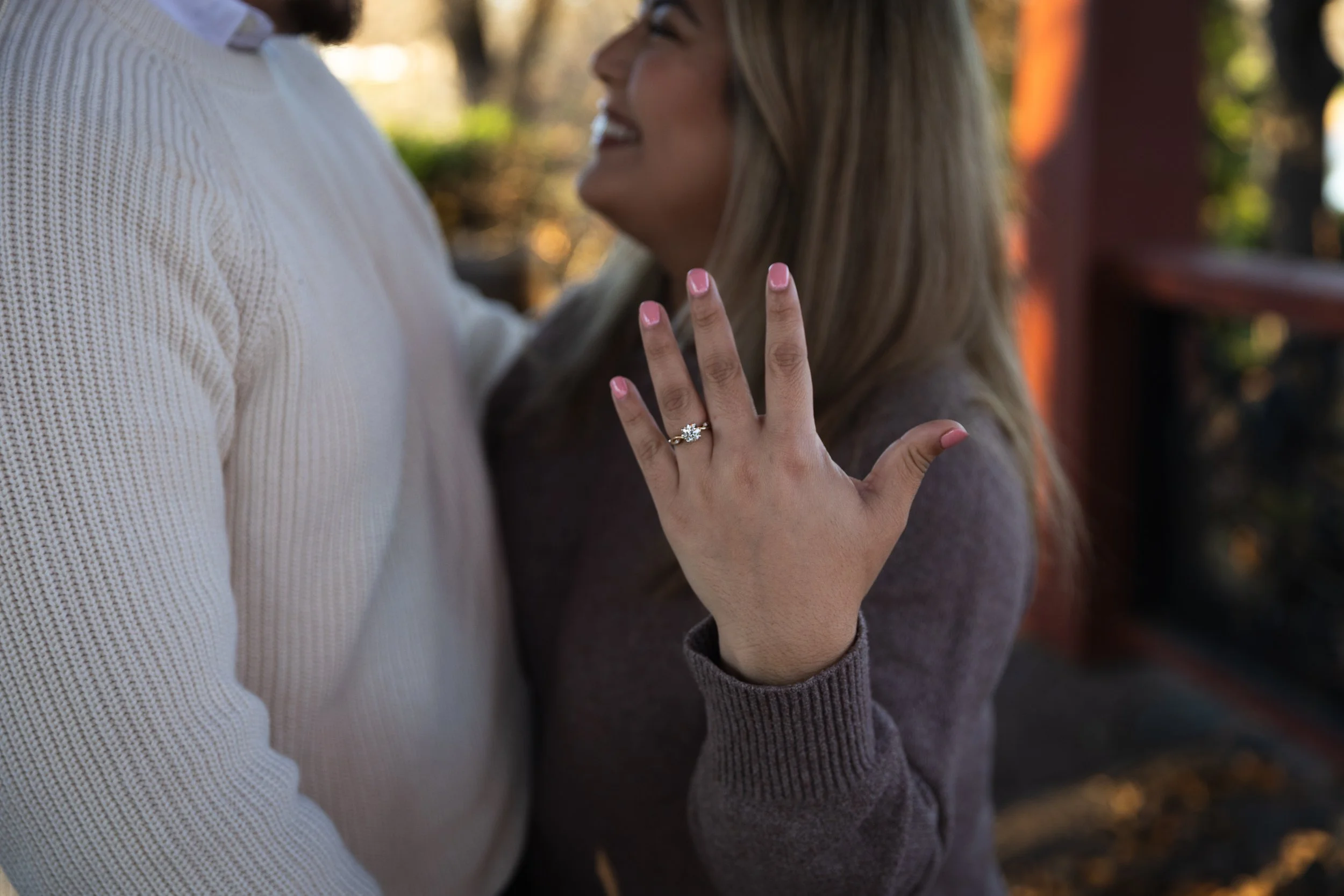 A woman with blonde hair wearing a gray sweater showing an engagement ring, closely embracing a man in a cream-colored sweater outdoors.