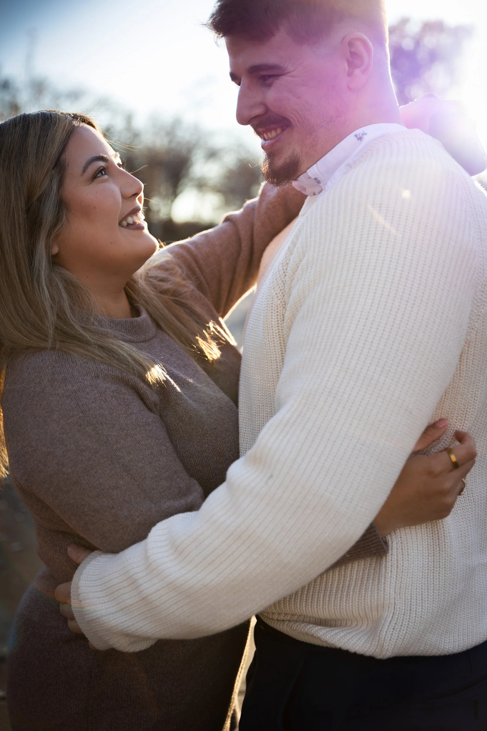 A man and woman are smiling and embracing outdoors with sunlight in the background.