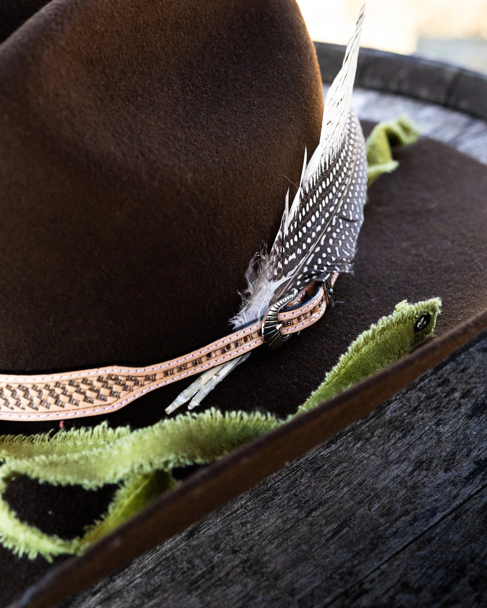 Close-up of a brown hat with a feather and a decorative band, resting on a dark wooden surface with green fabric trim in the foreground.