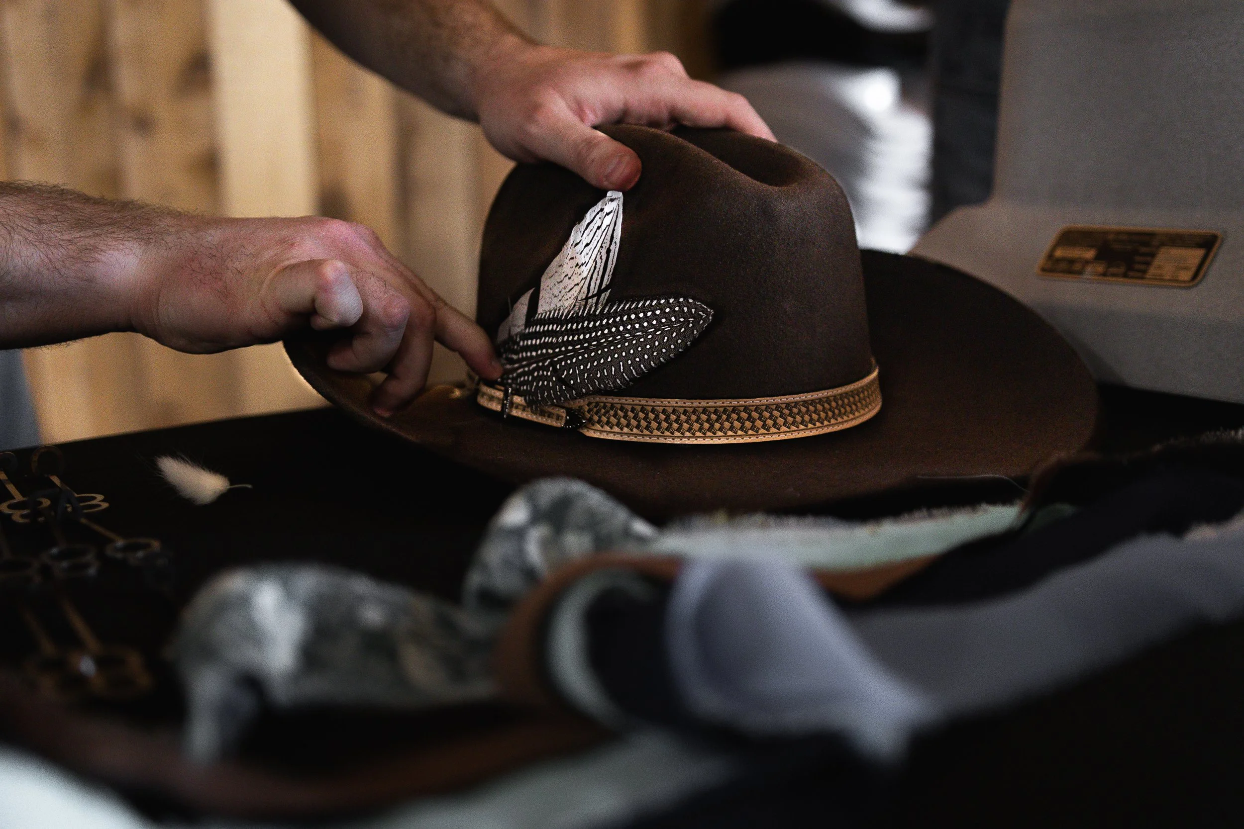 A person's hands adjusting a dark brown fedora with a decorative band, surrounded by hats and accessories on a table.