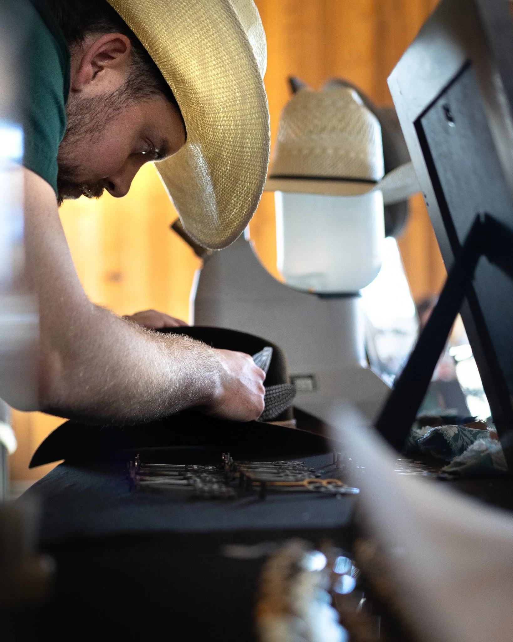 A person wearing a large straw hat leaning over a table, with a laptop, notes, and a coffee mug visible.
