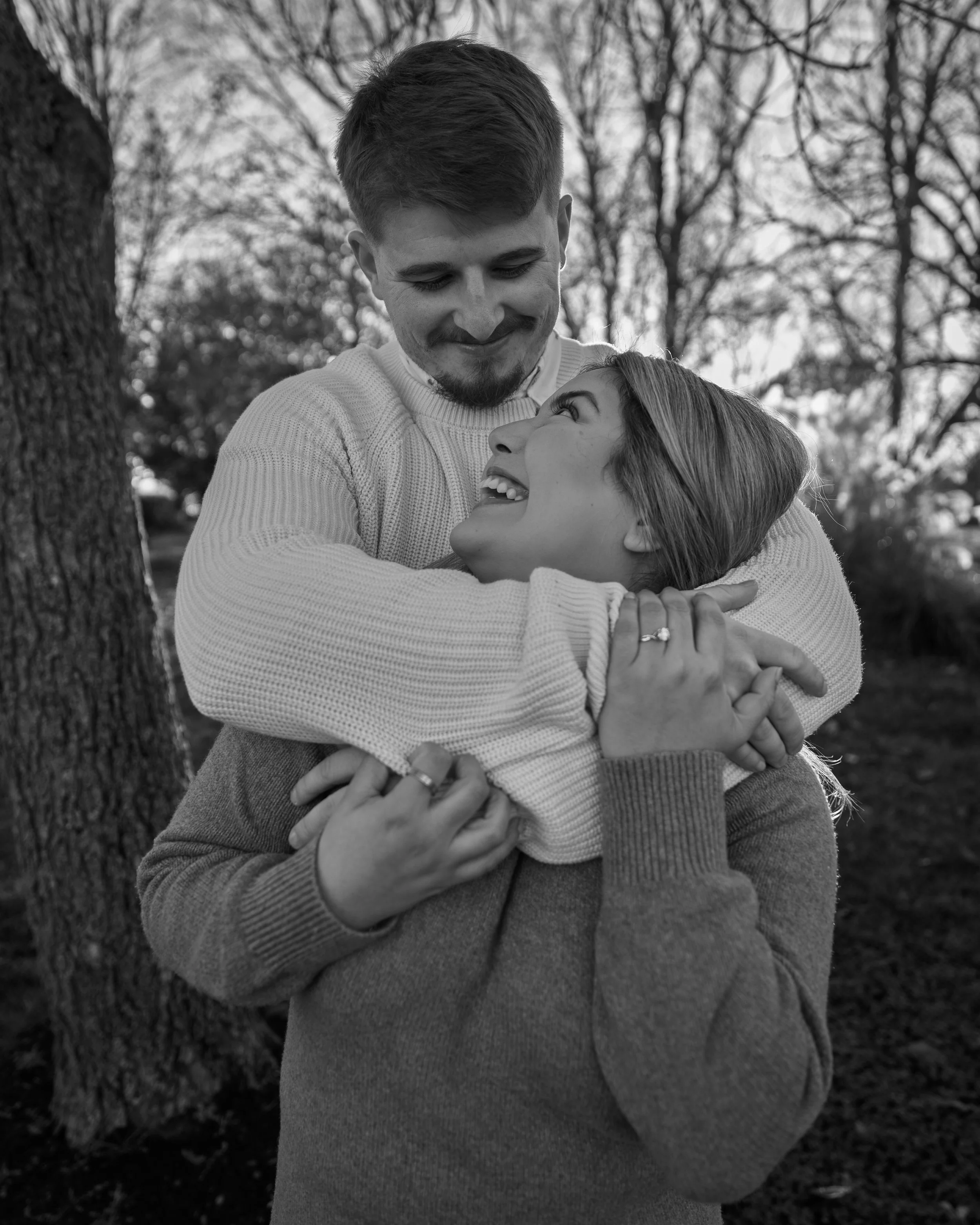 A black and white photo of a happy couple outdoors. The man is smiling and looking down, while the woman looks up at him with a joyful expression. They are embracing each other near a tree, with a background of leafless trees.