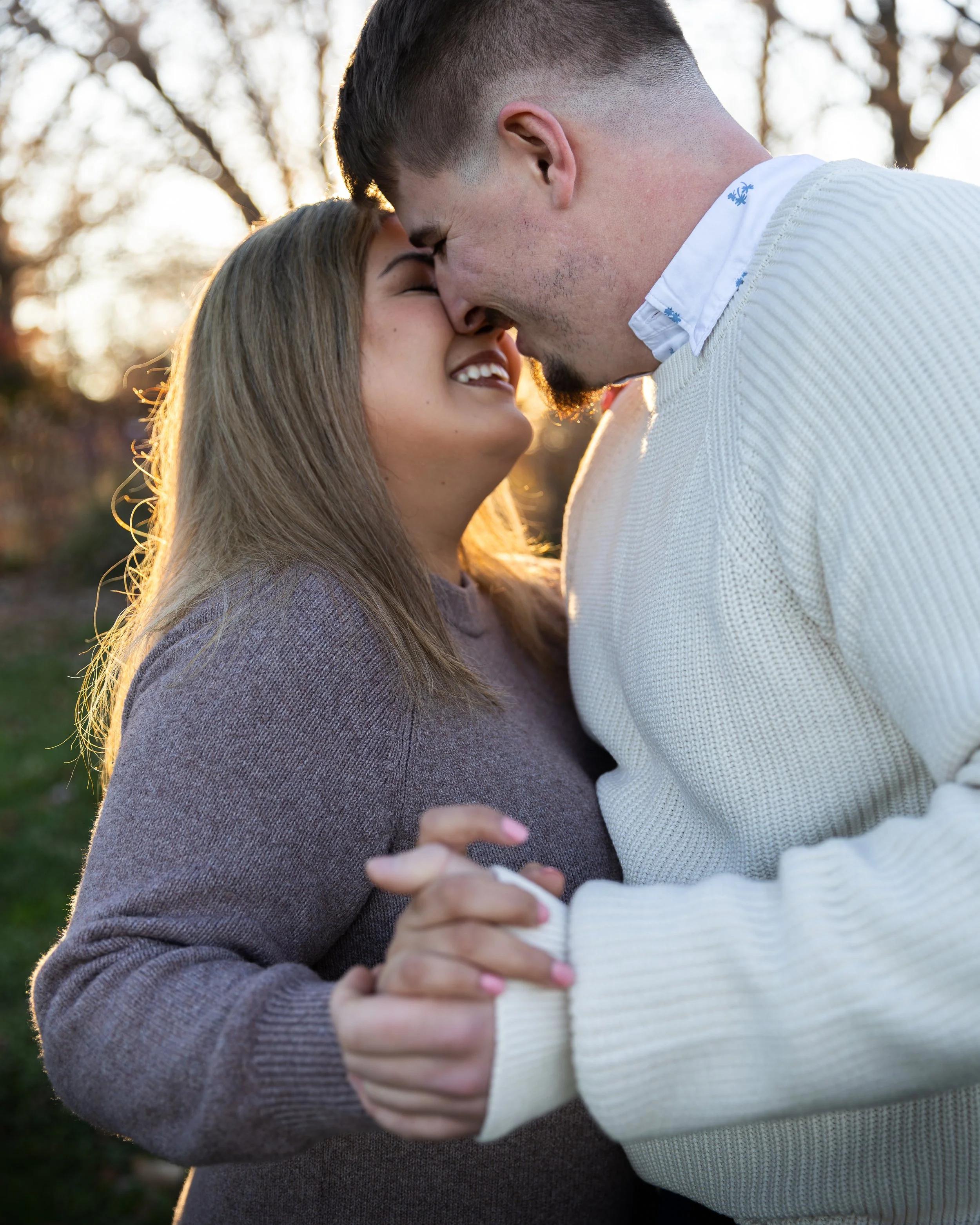 A couple embracing outdoors during sunset, smiling with faces close, in a park with blurred trees in the background.