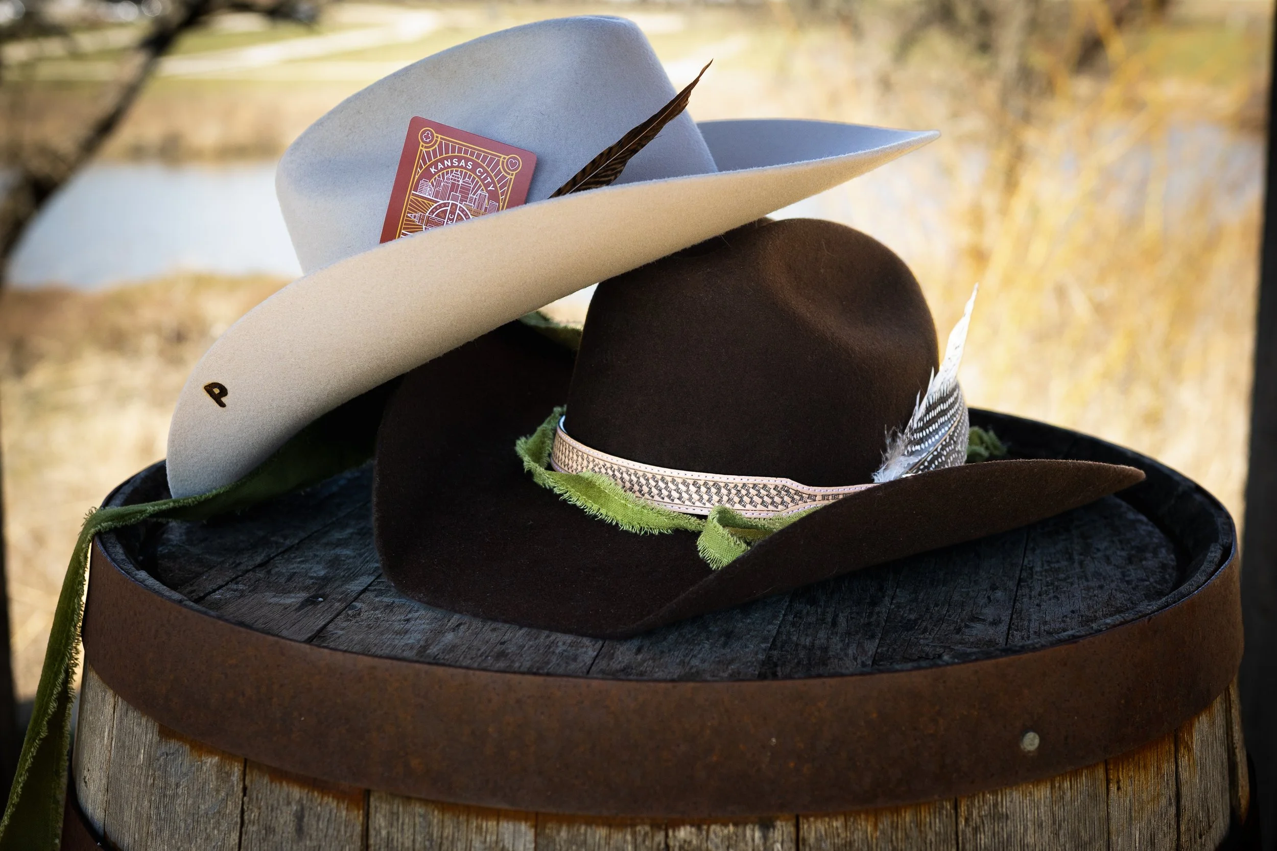 Various cowboy hats, including a white hat with a card and feather and a brown hat with a feather, are displayed on a circular wooden stand outdoors, with a blurred background of trees and grass.