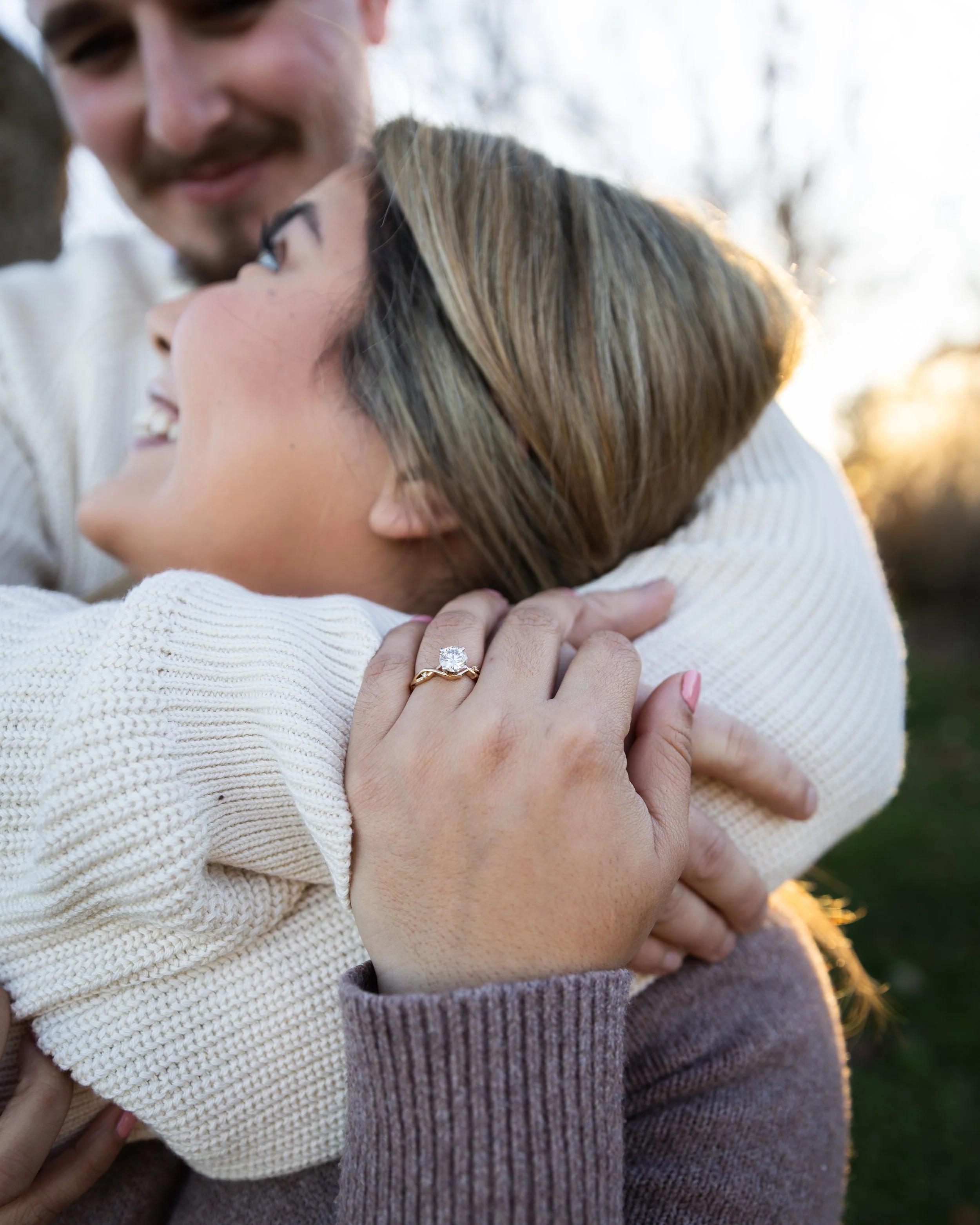 A couple hugging outdoors during sunset, with the woman's left hand showing an engagement ring. The man has facial hair and is smiling, while the woman has brown hair and is smiling, looking happy.