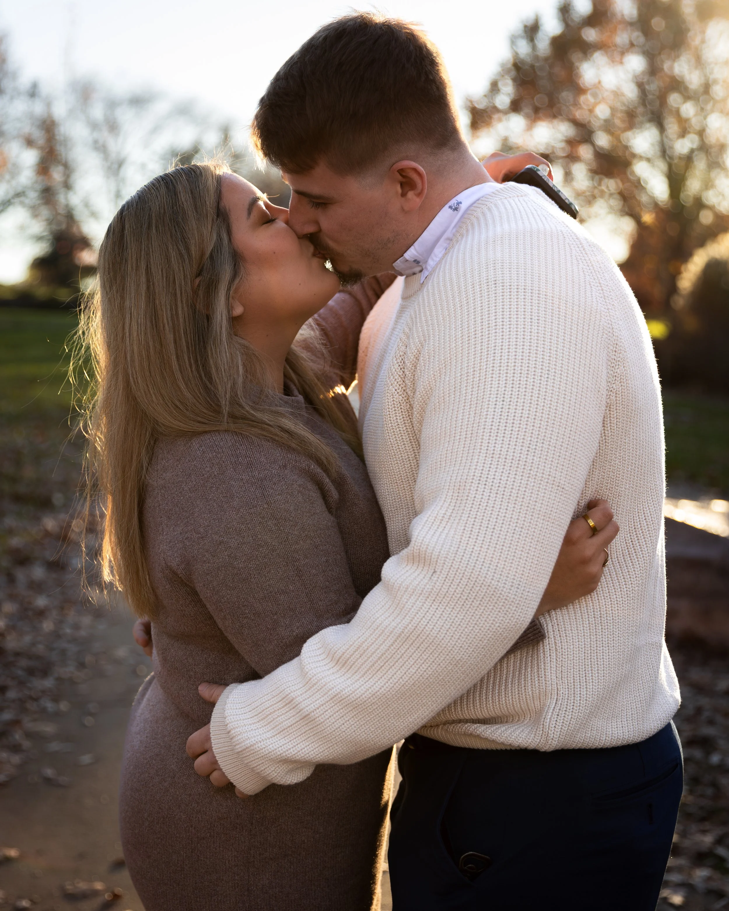 A couple kissing outdoors during the golden hour, with blurred trees in the background.