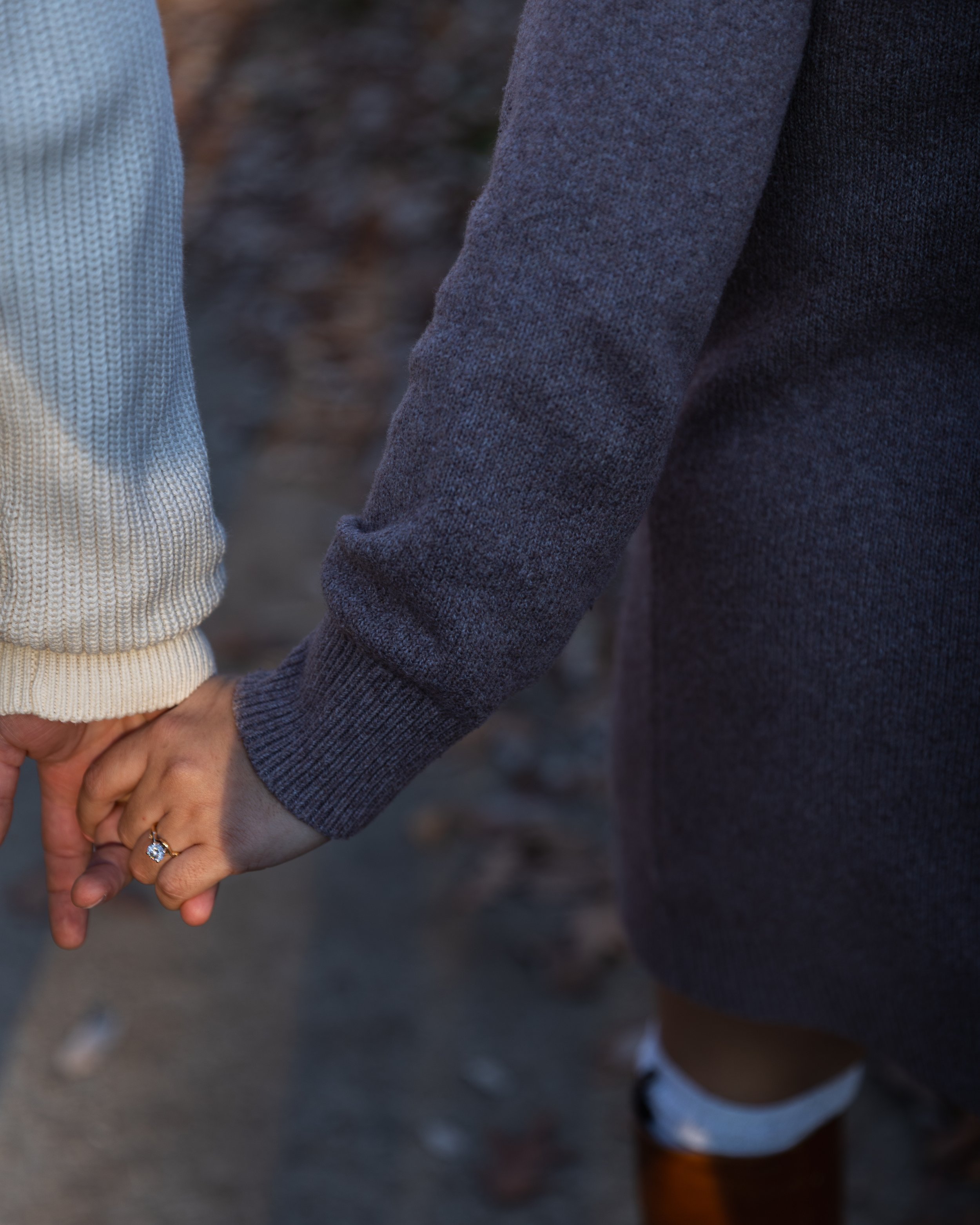 Close-up of two people holding hands, one wearing a gray sweater and the other wearing a cream knit sweater, with a blurred outdoor background.