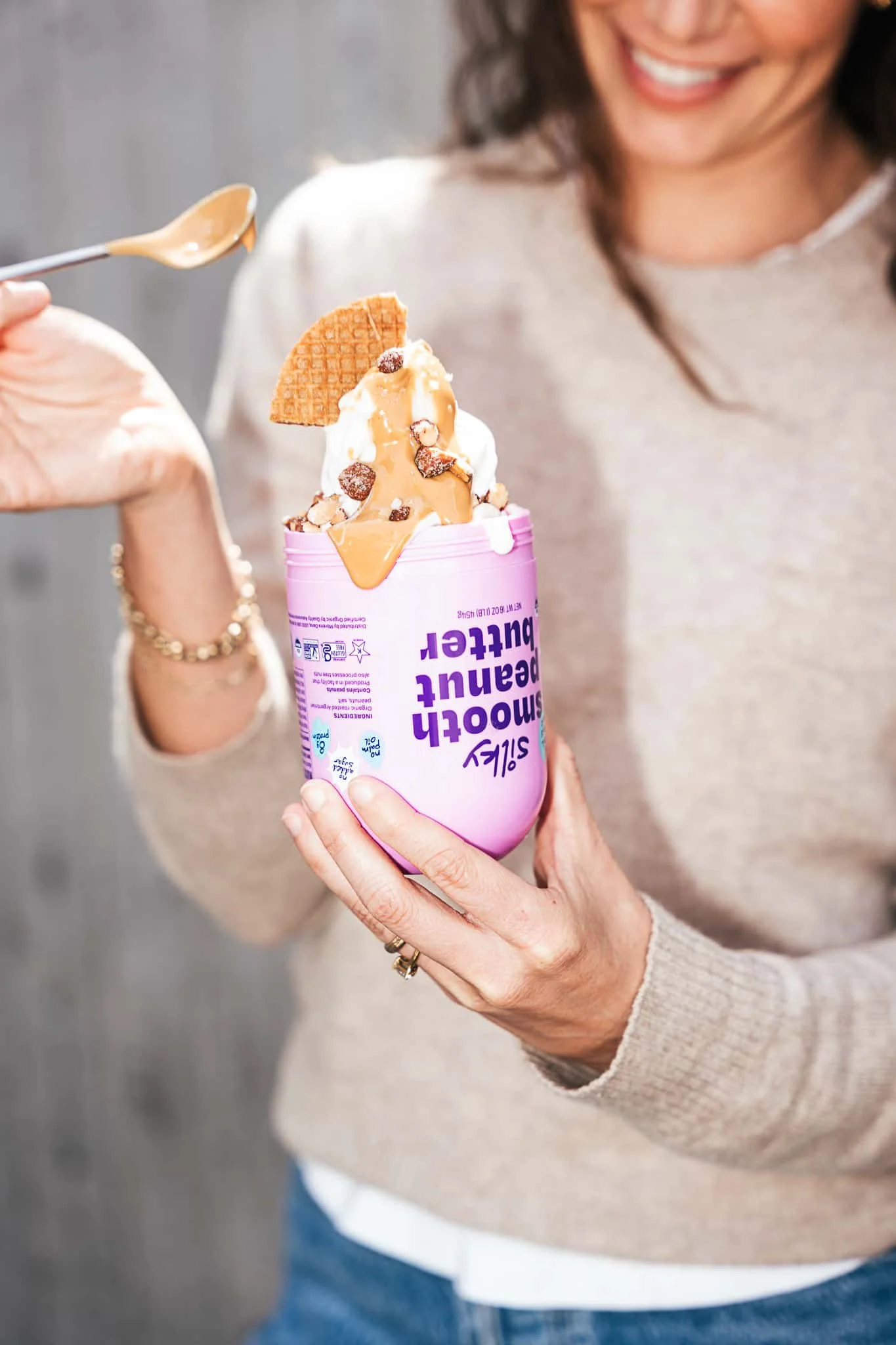 A woman holding a pink container of frozen yogurt with caramel sauce and chocolate pieces, smiling, while using a spoon to add more caramel to the ice cream.