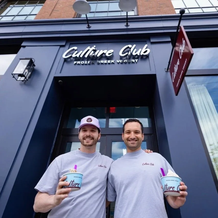 Two smiling men standing outside a "Culture Club Frozen Greek Yogurt" shop, each holding a cup of frozen yogurt.