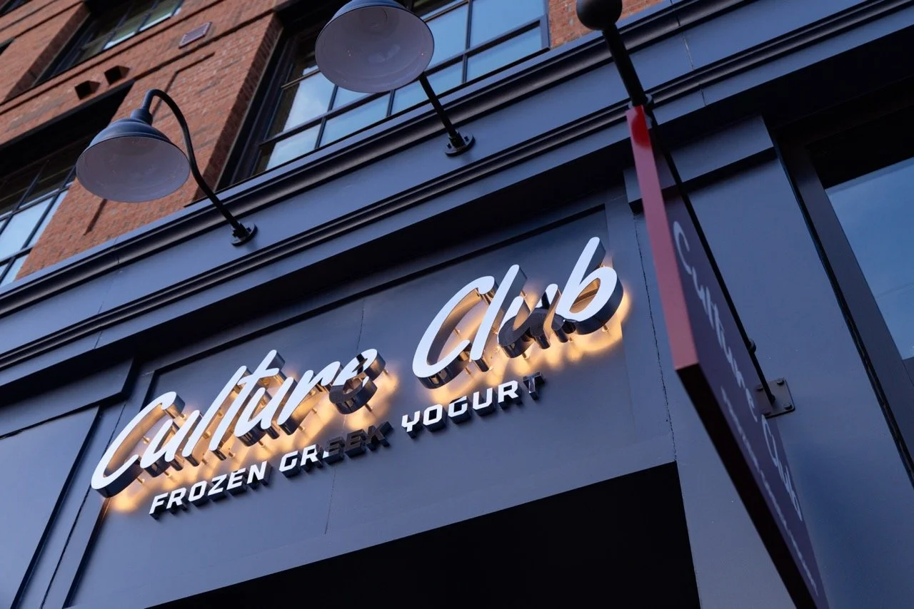 Outside view of a storefront sign for Culture Club frozen Greek yogurt with modern lighting fixtures above the sign and a brick building facade in the background.