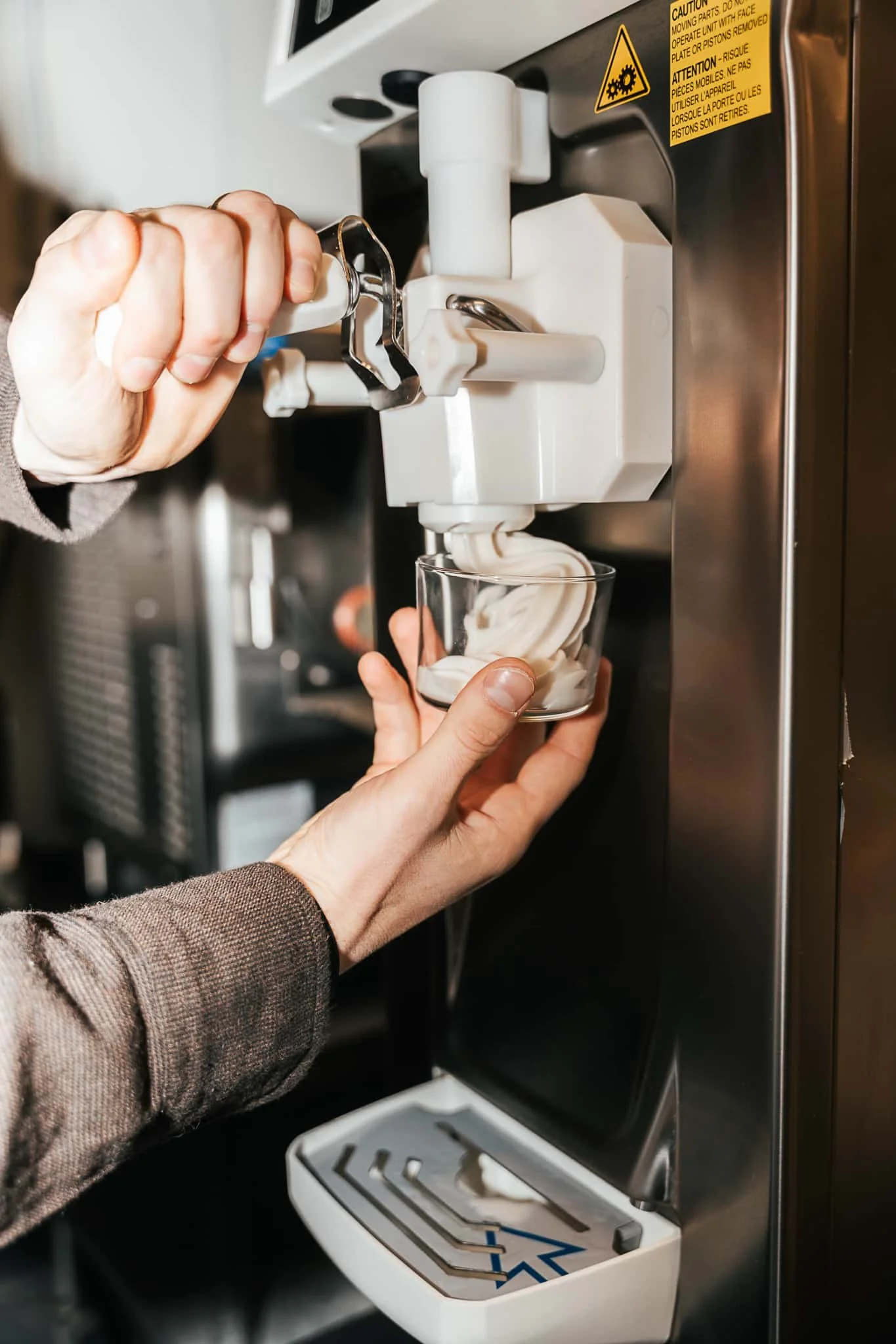 A person using a soft serve frozen yogurt machine to fill a glass with vanilla frozen yogurt.