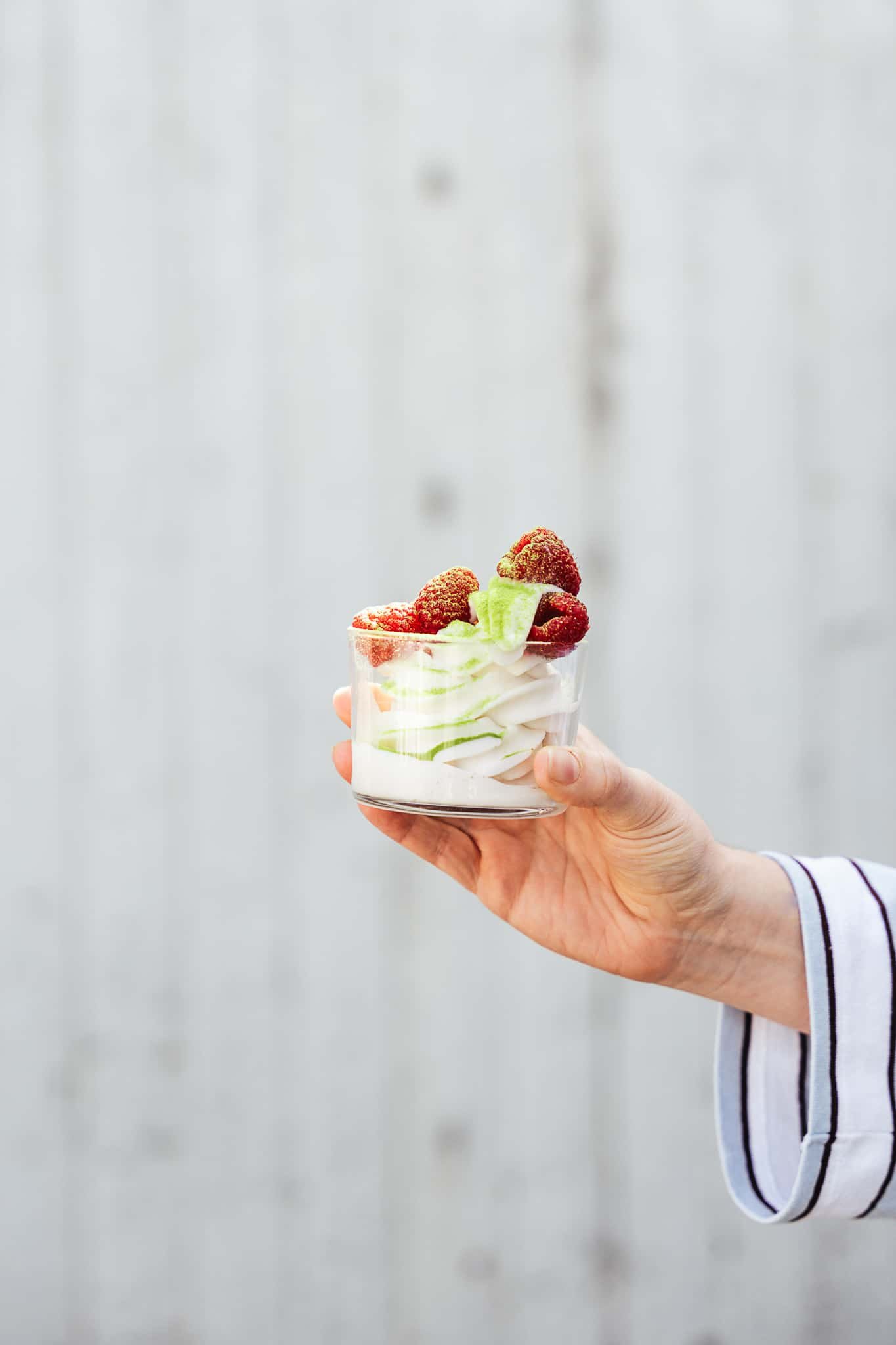 A hand holding a glass of frozen yogurt topped with raspberries and a green matcha powder against a plain, light-colored background. 