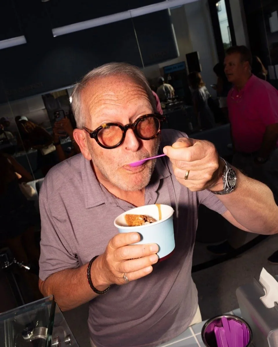 An older man wearing tortoiseshell glasses tastes a spoonful of frozen yogurt from a light blue cup.