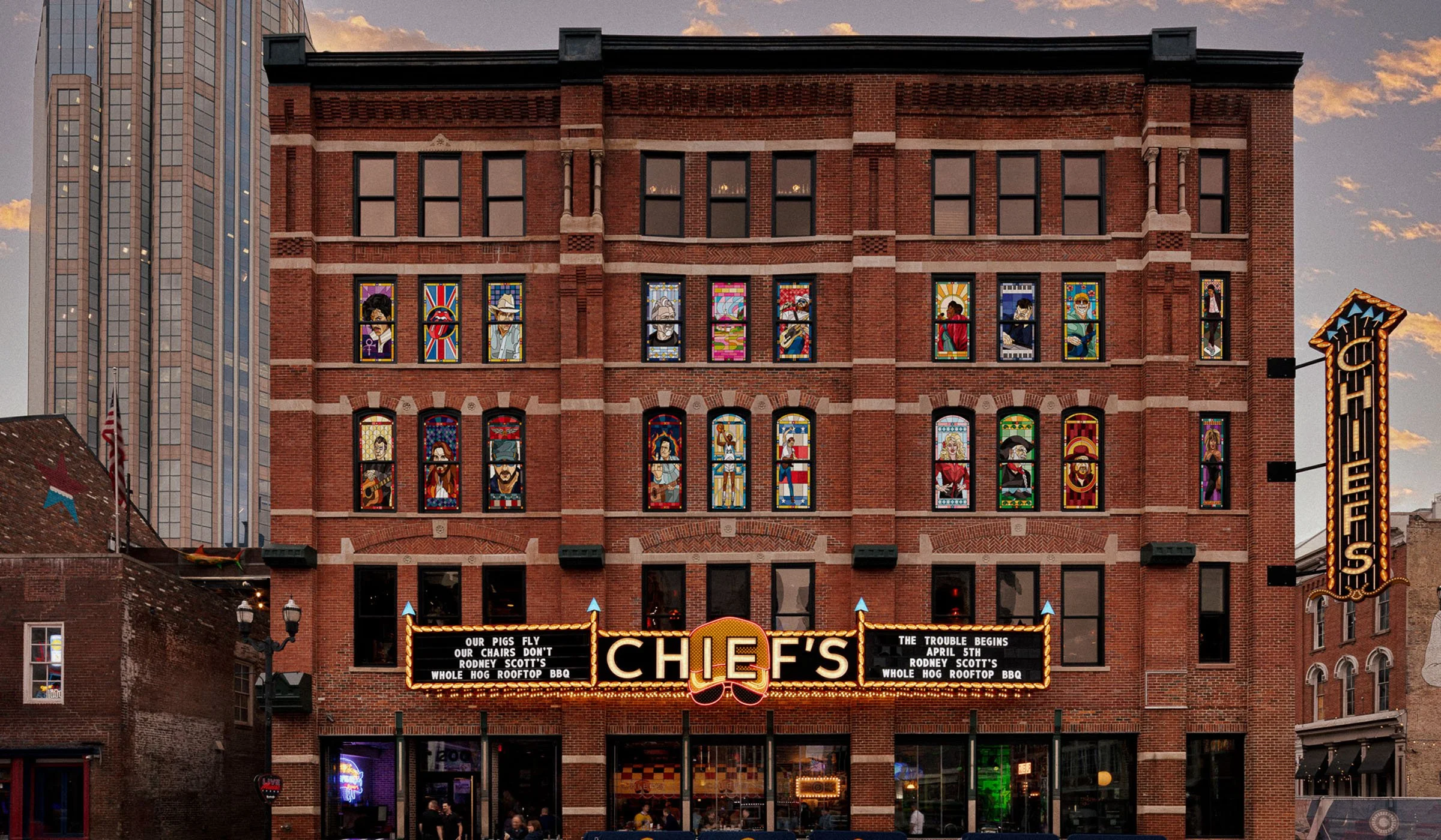 The image shows a multi-story brick building with colorful stained-glass windows depicting various characters and scenes, with a marquee sign and a theater or music venue entrance below. The sky in the background indicates it is during sunset or early evening.
