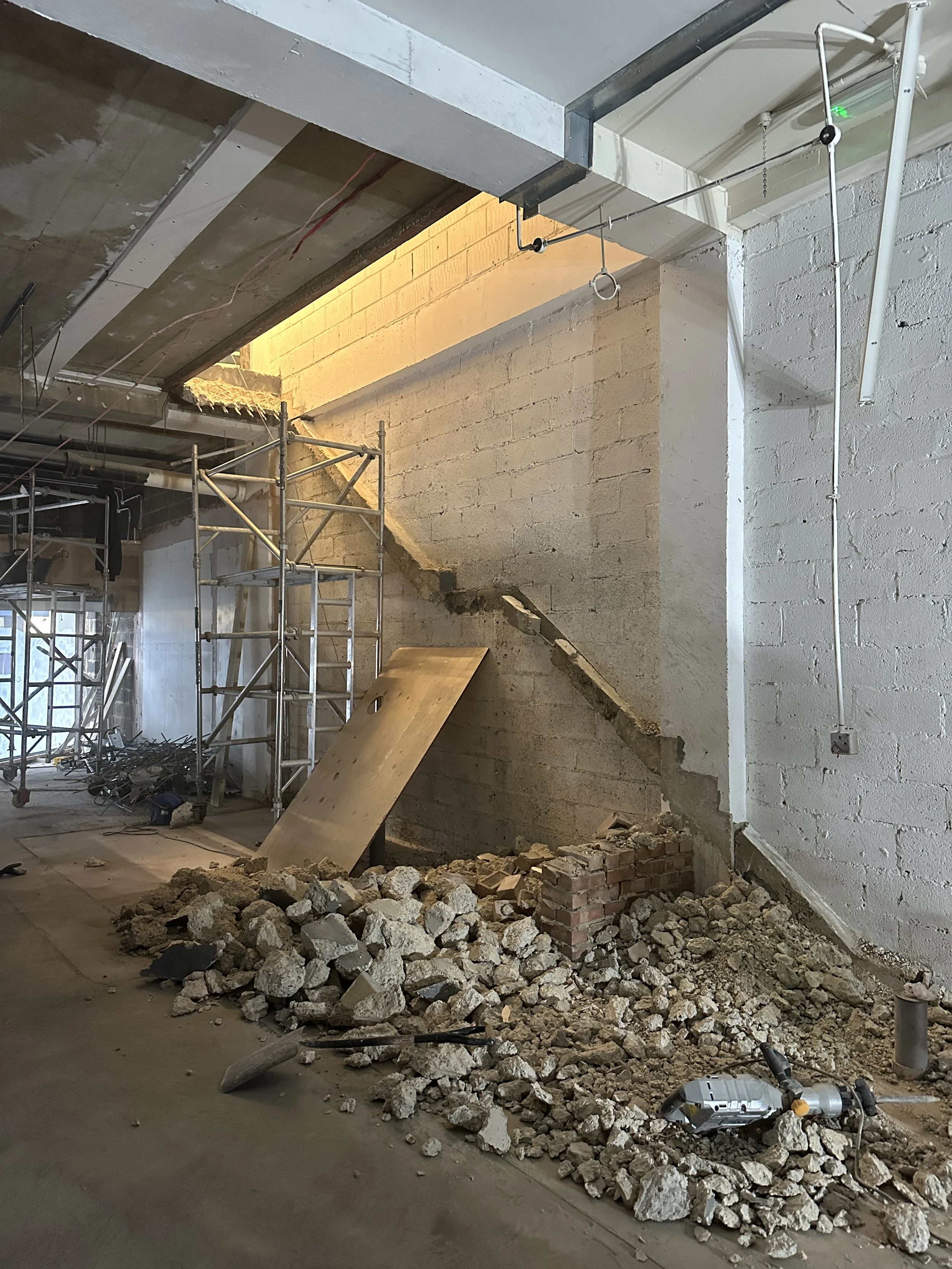 Construction site with scattered debris, rubble, and tools, including a saw, on a concrete floor. Scaffolding is in the background, and part of a brick wall is visible.