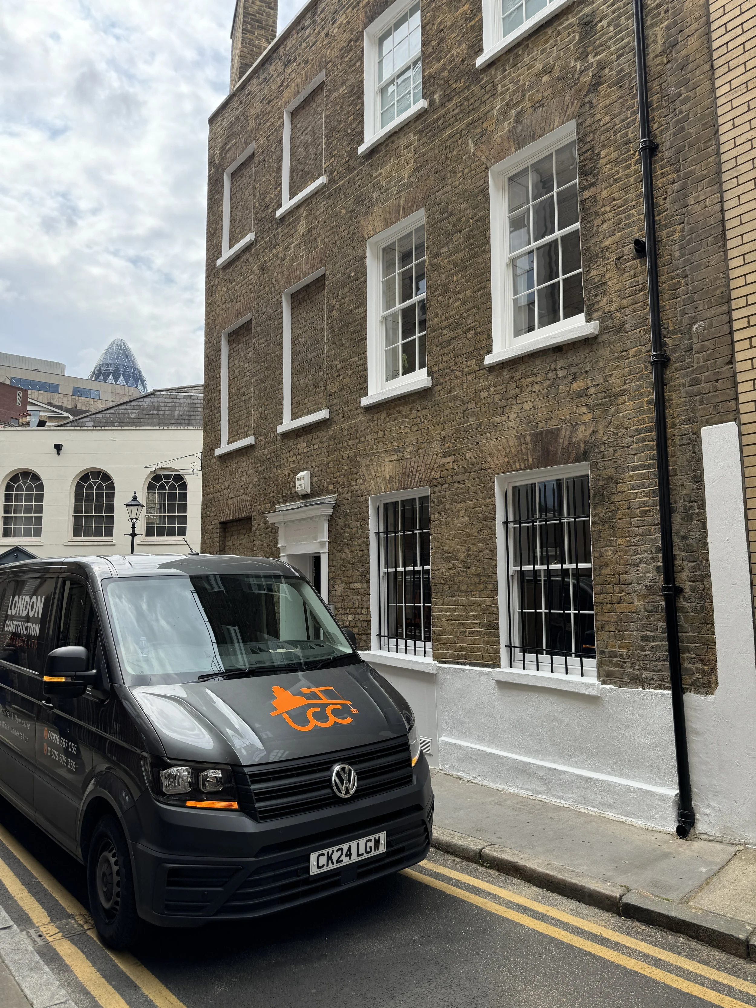 A black Volkswagen van parked on a city street beside a brick building with white-framed windows and black bars on the lower windows. The van has an orange logo and text on the hood and sides, with contact information. In the background, there are other buildings and a partly cloudy sky.