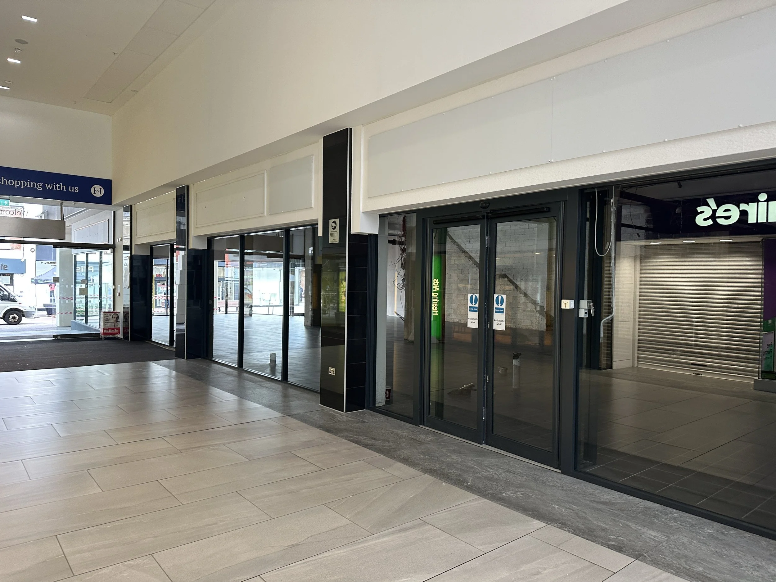 Empty shopping mall entrance with glass doors, shuttered store, and a sign indicating shopping with us.
