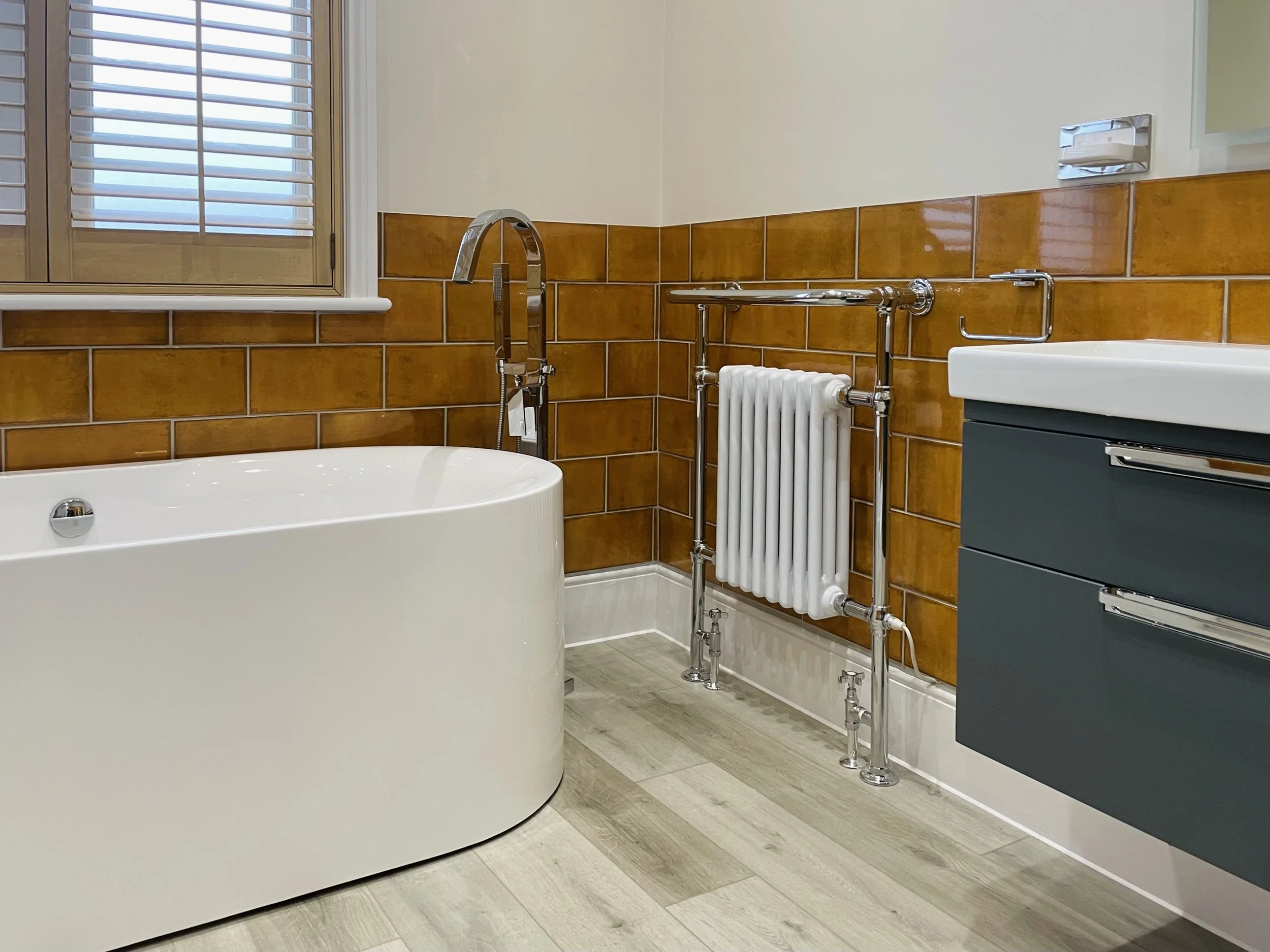 A modern bathroom with a bathtub, brown tiled walls, a radiator, a faucet, a soap dish, a mirror, and a window with shutters.