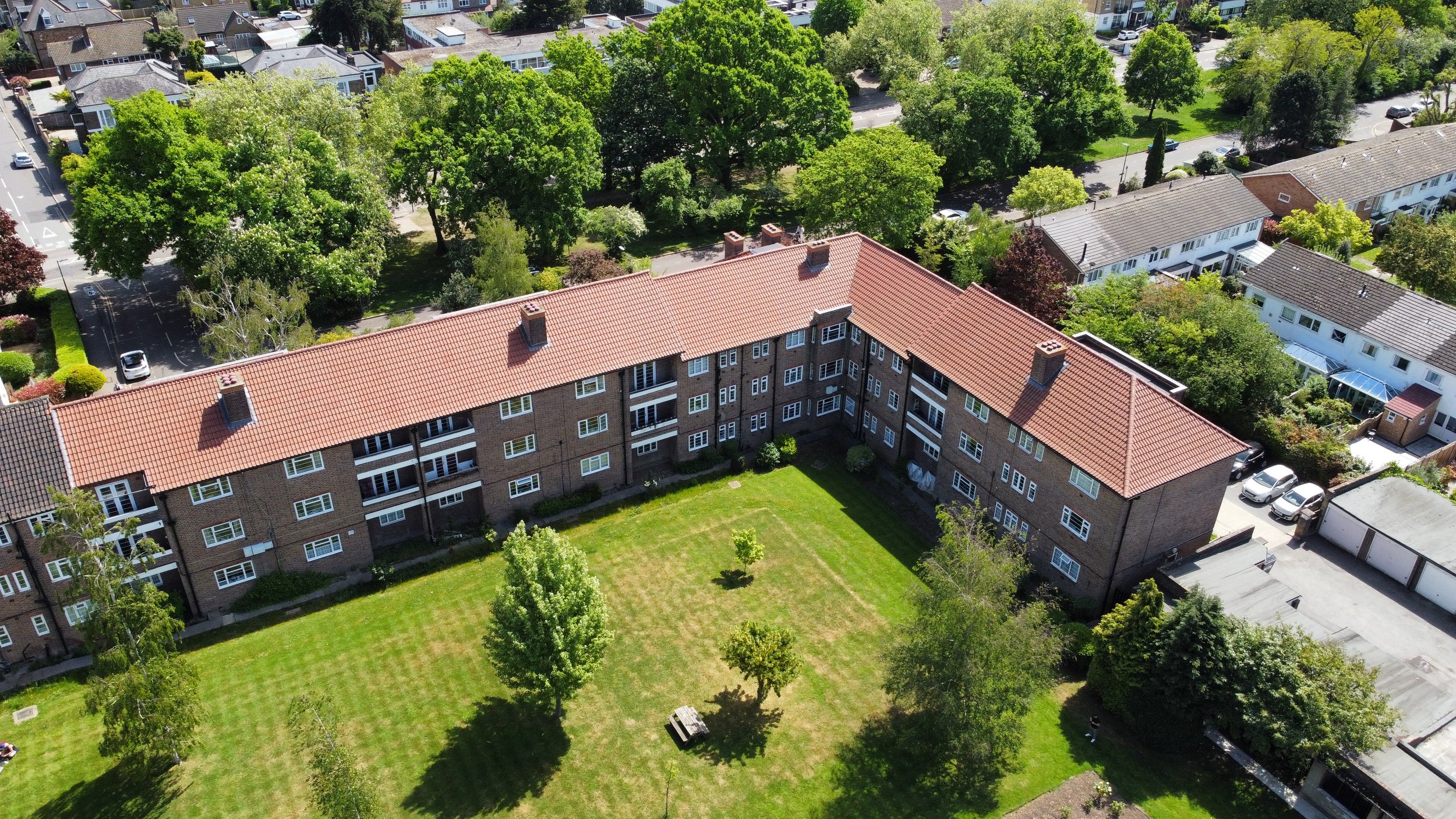 Aerial view of a residential area showing a large brick apartment building with a red-tiled roof, surrounding green trees, lawns, parking lots, and neighboring houses.