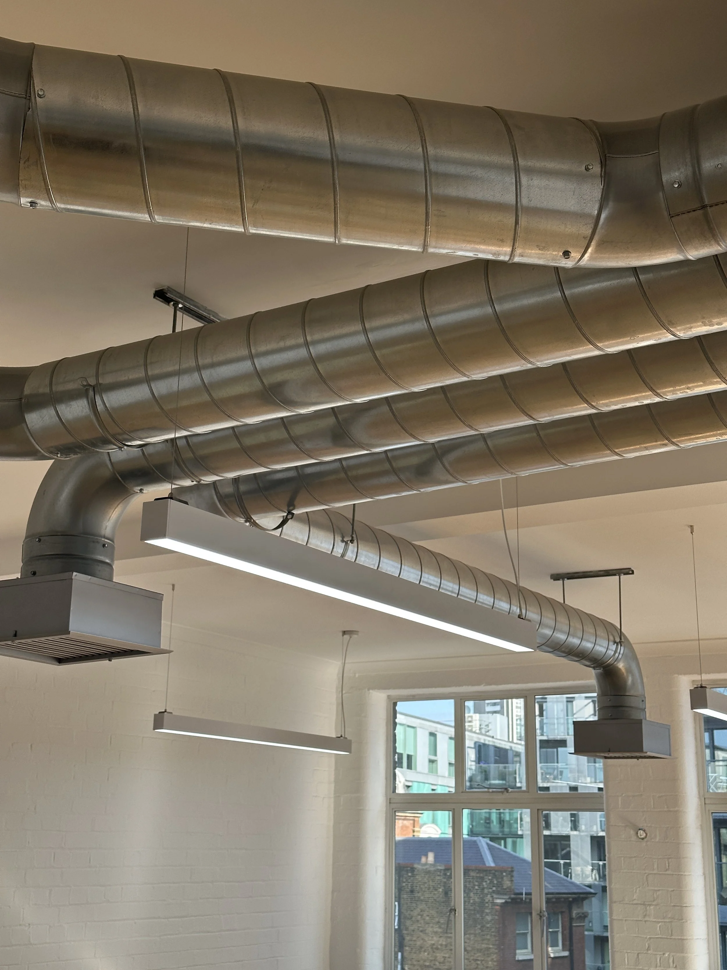 Indoor space with exposed metallic ductwork on ceiling, white brick walls, and large windows showing buildings outside.
