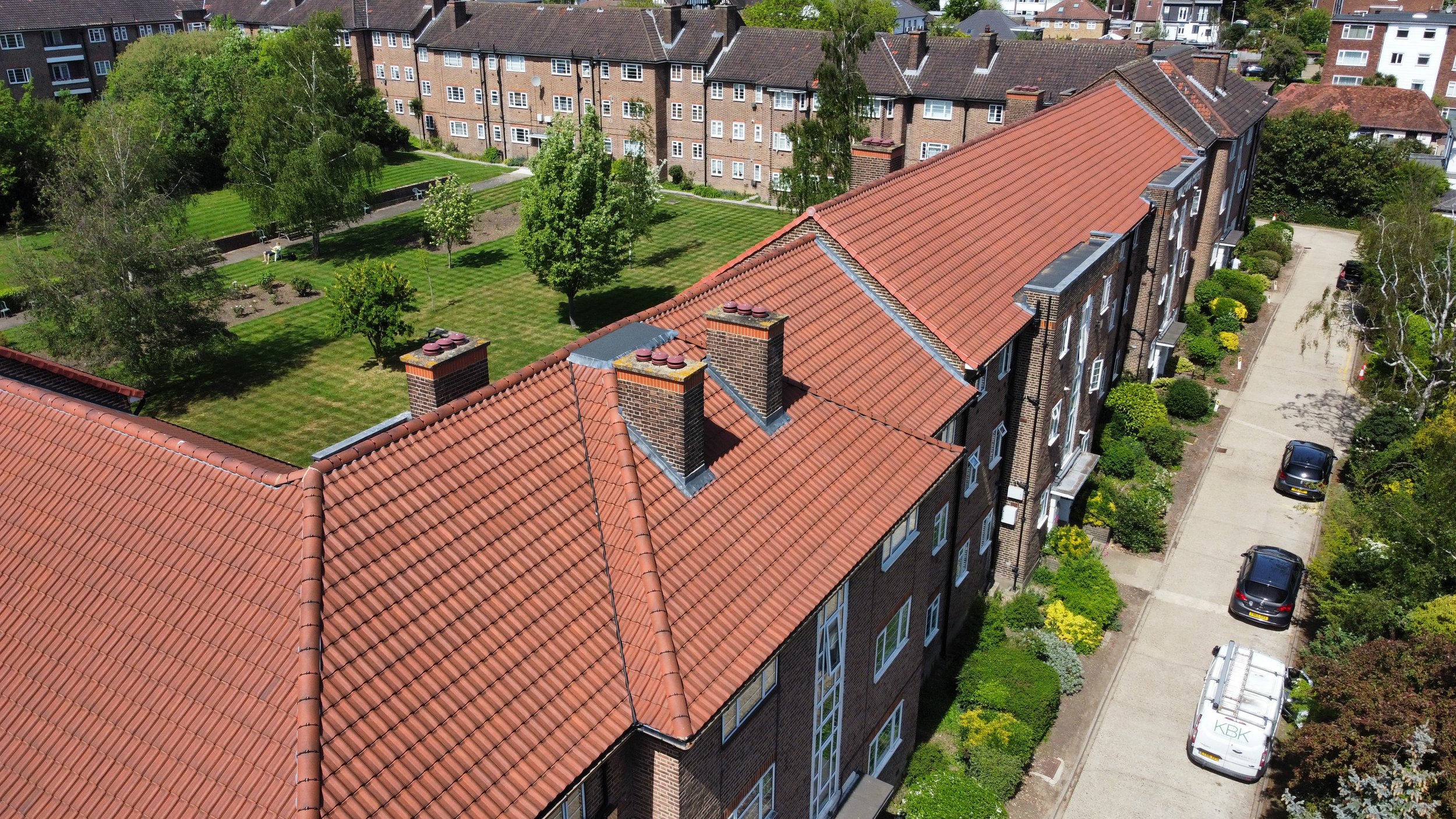 An aerial view of a residential complex with red-tiled roofs, a green park with trees, benches, and walking paths, and a parking lot with cars. Tall apartment buildings surround the park.