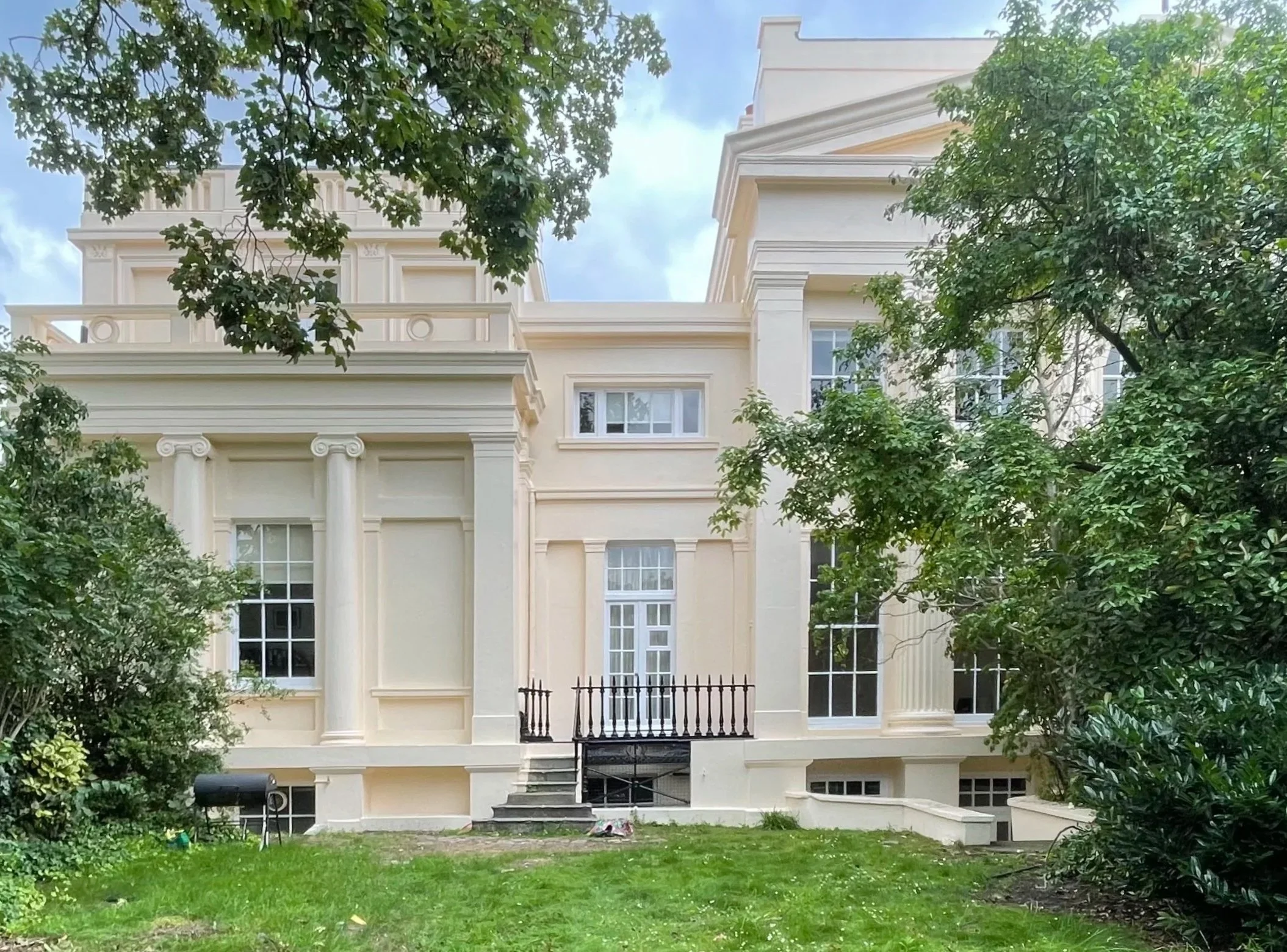 A white, multi-story house with classical architectural details, large windows, and a small front yard with green grass, surrounded by trees.