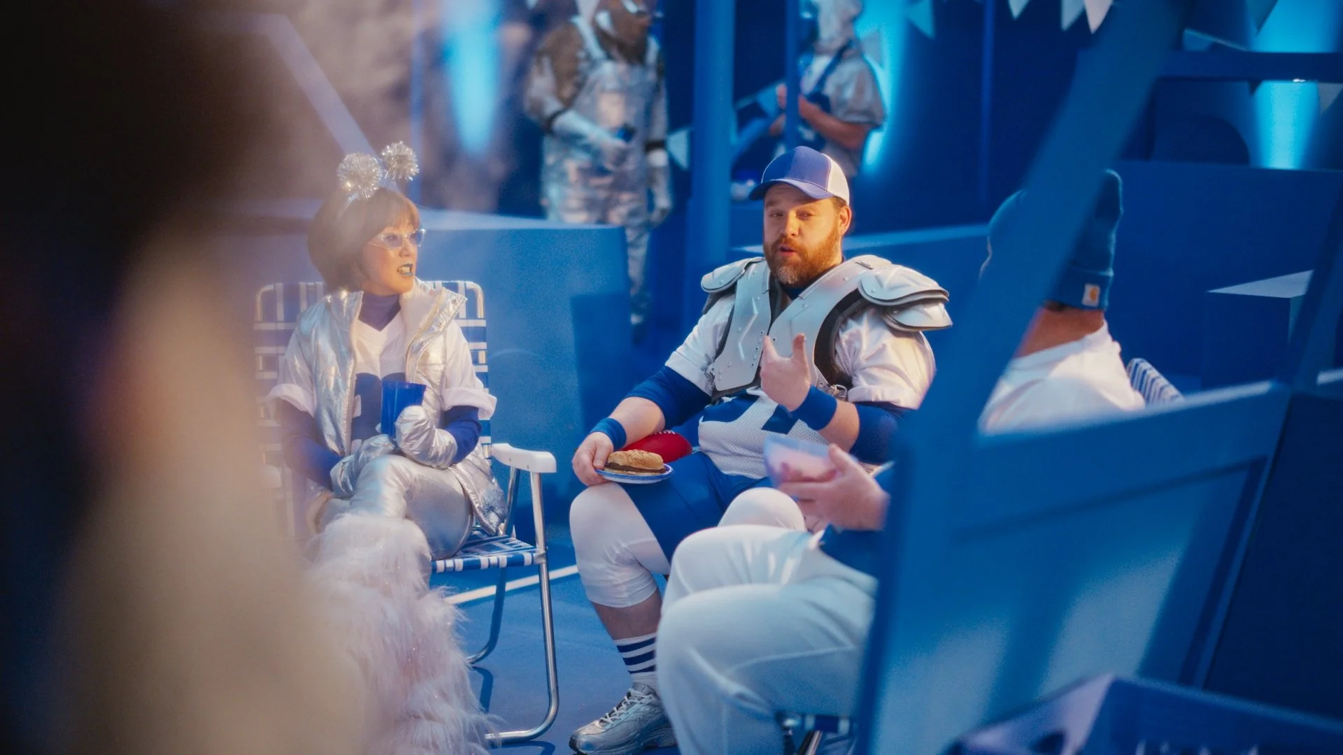 People in athletic costumes on a set with blue lighting, two sitting in chairs, one with a plate of food, the other holding a drink.