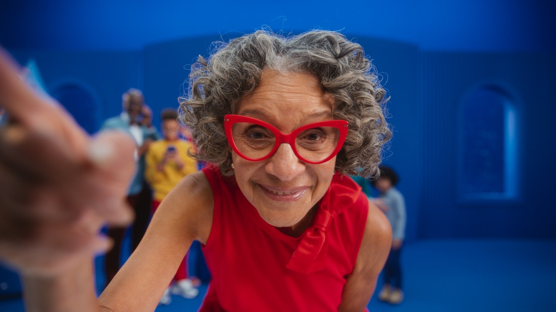 Close-up of an elderly woman with curly gray hair wearing large red glasses and a red sleeveless dress, smiling and leaning toward the camera with a playful expression in a blue room with children in the background.